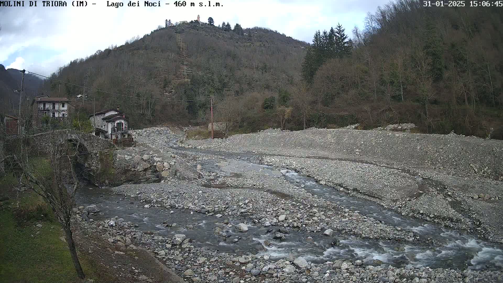 A rocky river flows through a valley under a partly cloudy sky, past a stone bridge and small buildings nestled against a hillside of leafless trees.