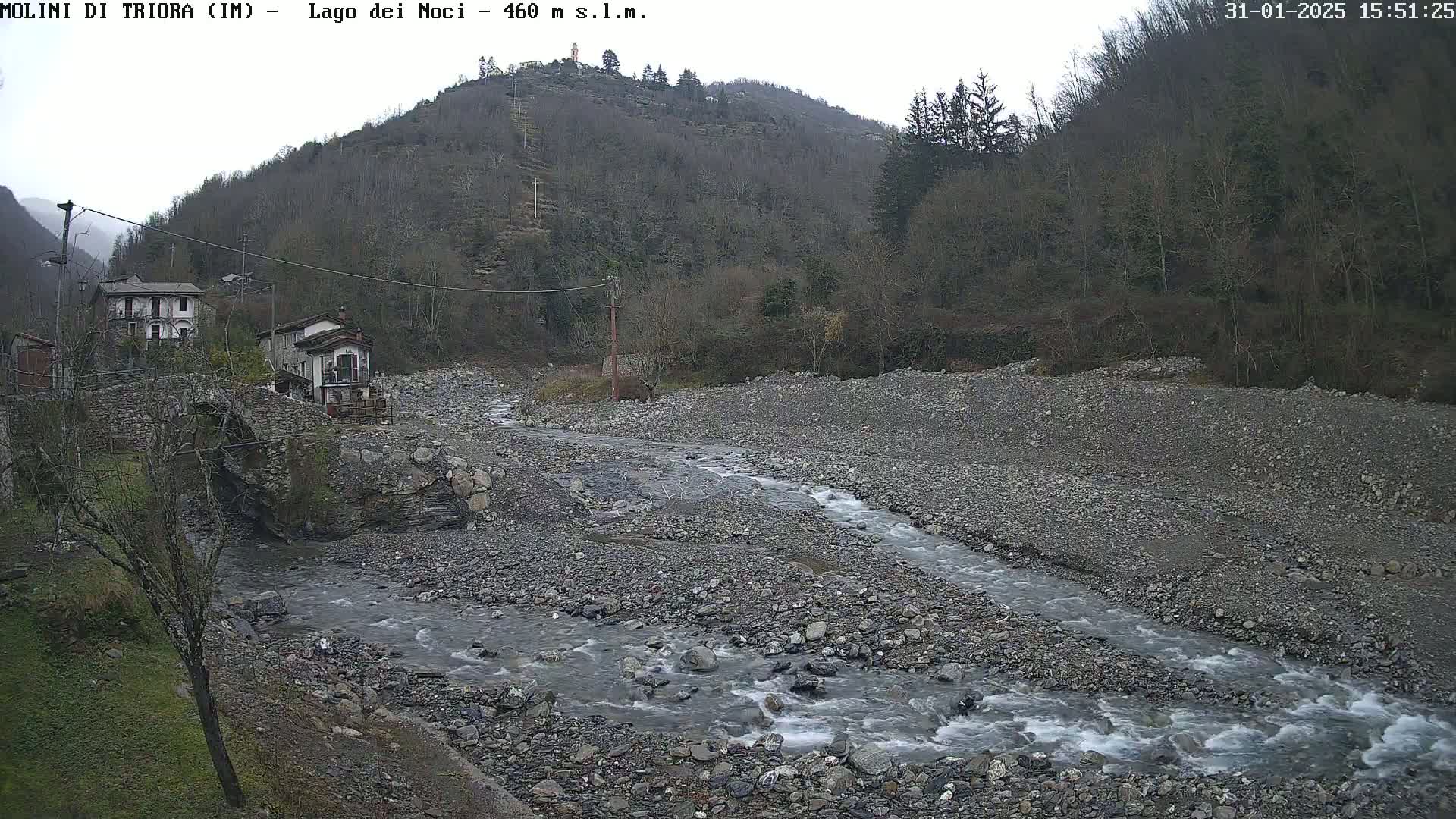 A rocky, gray river flows through a valley under an overcast sky, past a stone bridge and a small village nestled against a hillside of bare trees.