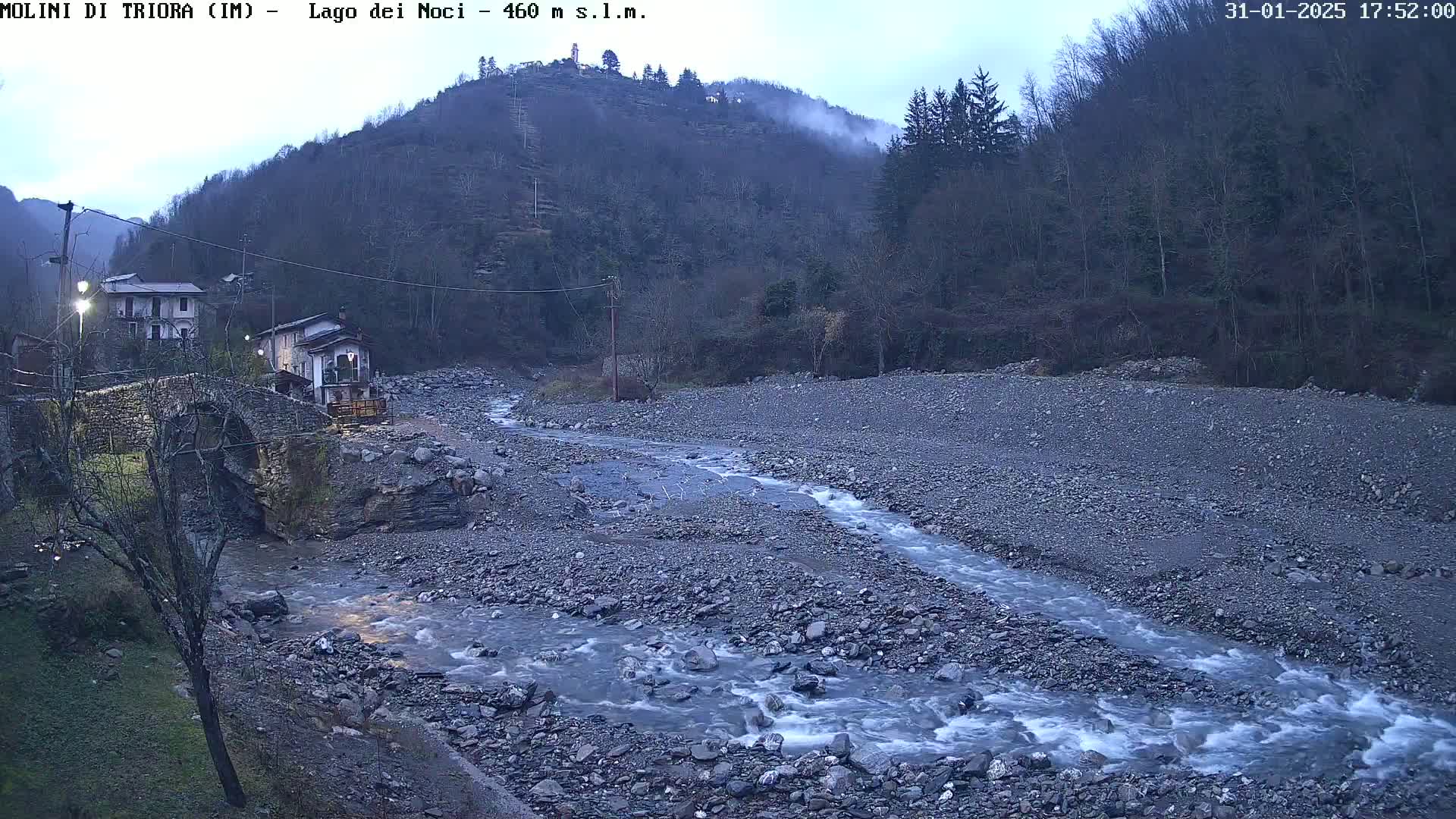 A rocky river flows through a valley under an overcast sky, passing under a stone bridge near some buildings nestled at the foot of a forested hill.