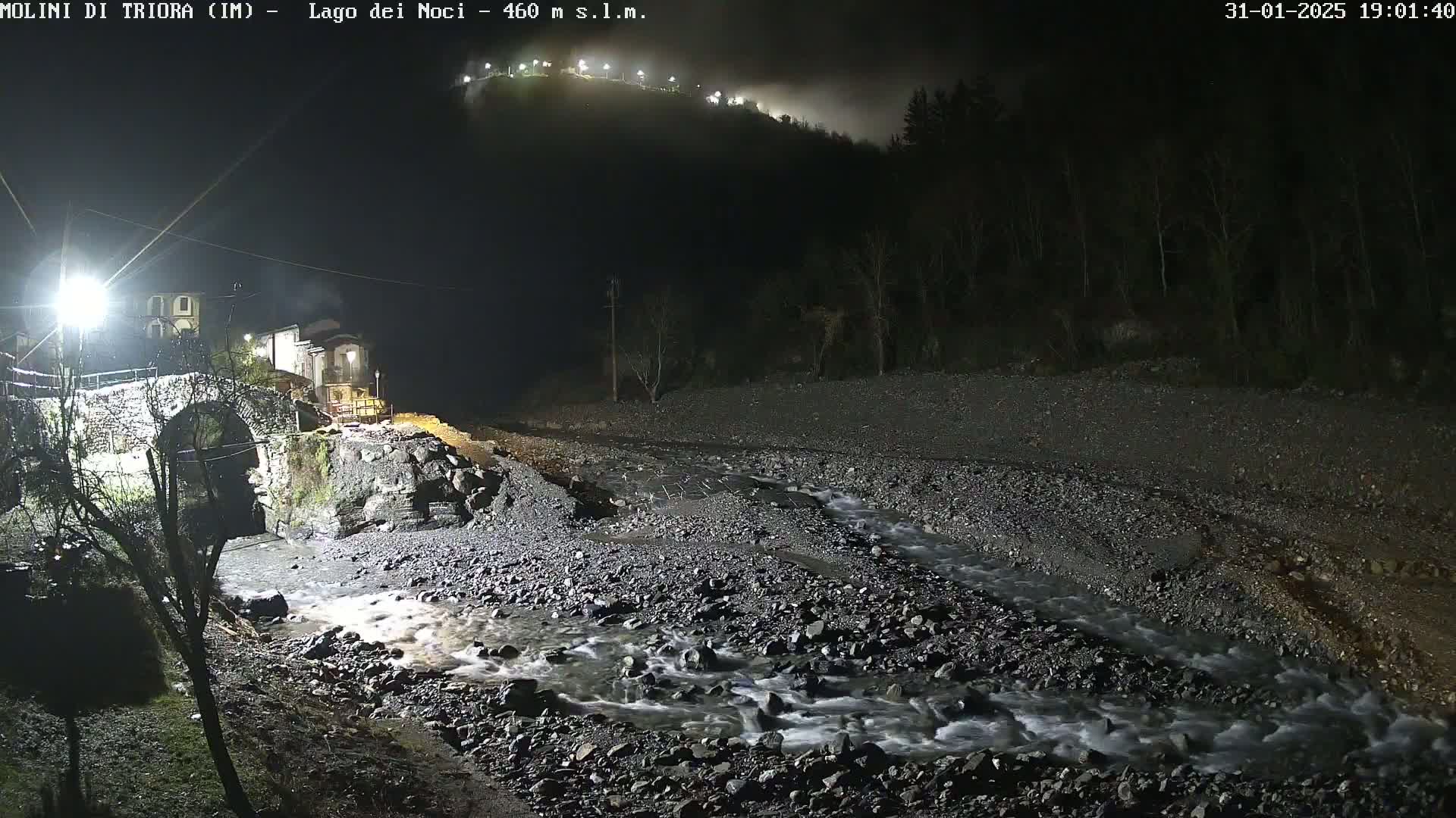 A nighttime view of a rocky riverbed with a small stream flowing through it, near a stone bridge and village under a foggy sky illuminated by artificial lights.