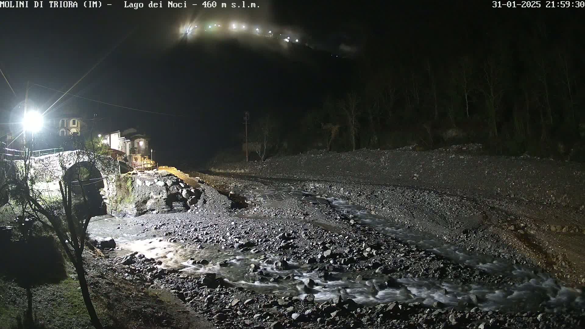 A rocky river flows under a stone bridge near a small village at night, under a mostly clear sky.