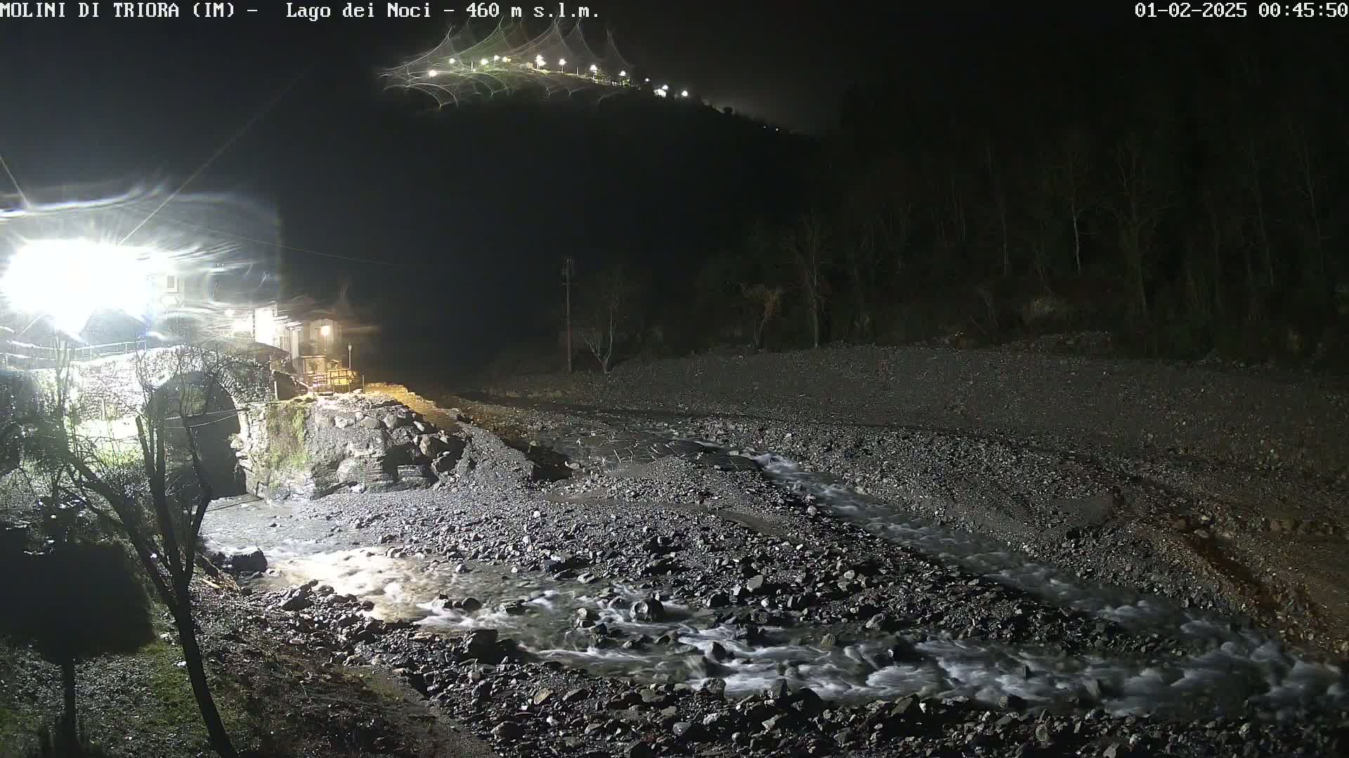 A nighttime view of a rocky stream flowing under a stone bridge near a village, illuminated by artificial light.