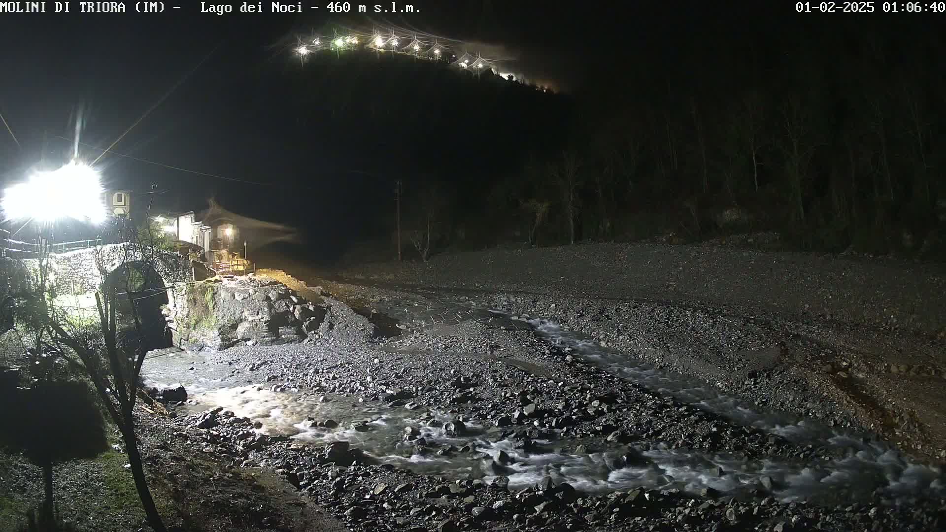 At night, a rocky stream flows past a stone arch bridge near a village, illuminated by artificial lights.