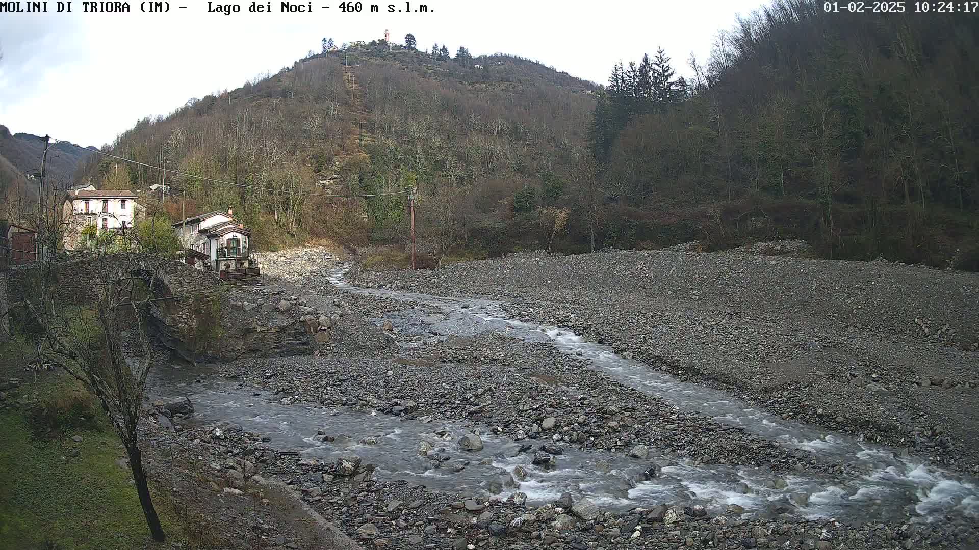 A rocky stream flows through a valley, past a small village nestled beside a hill under an overcast sky.