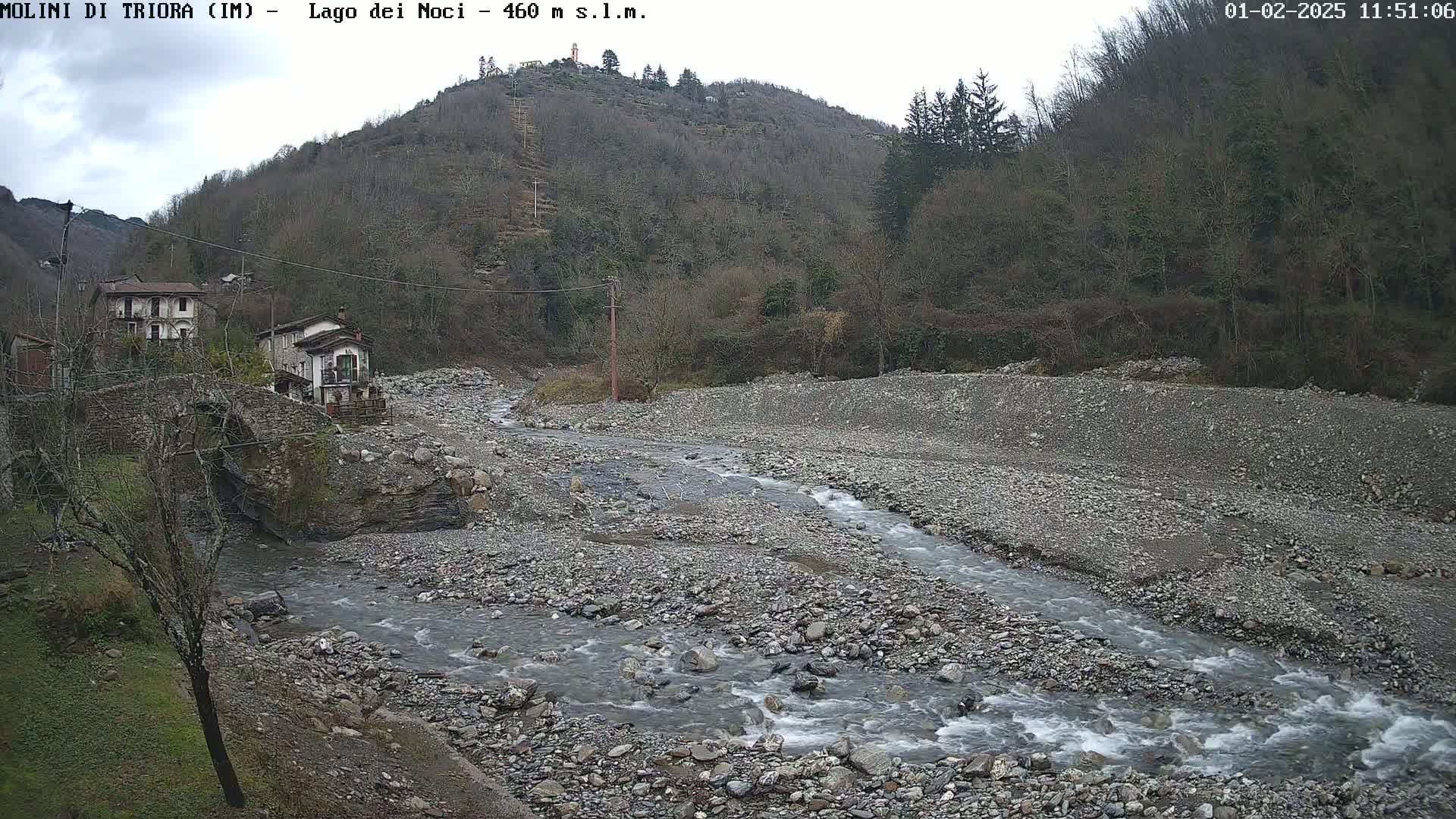 A rocky river flows through a valley under an overcast sky, past a small village nestled against a hillside covered in leafless trees.