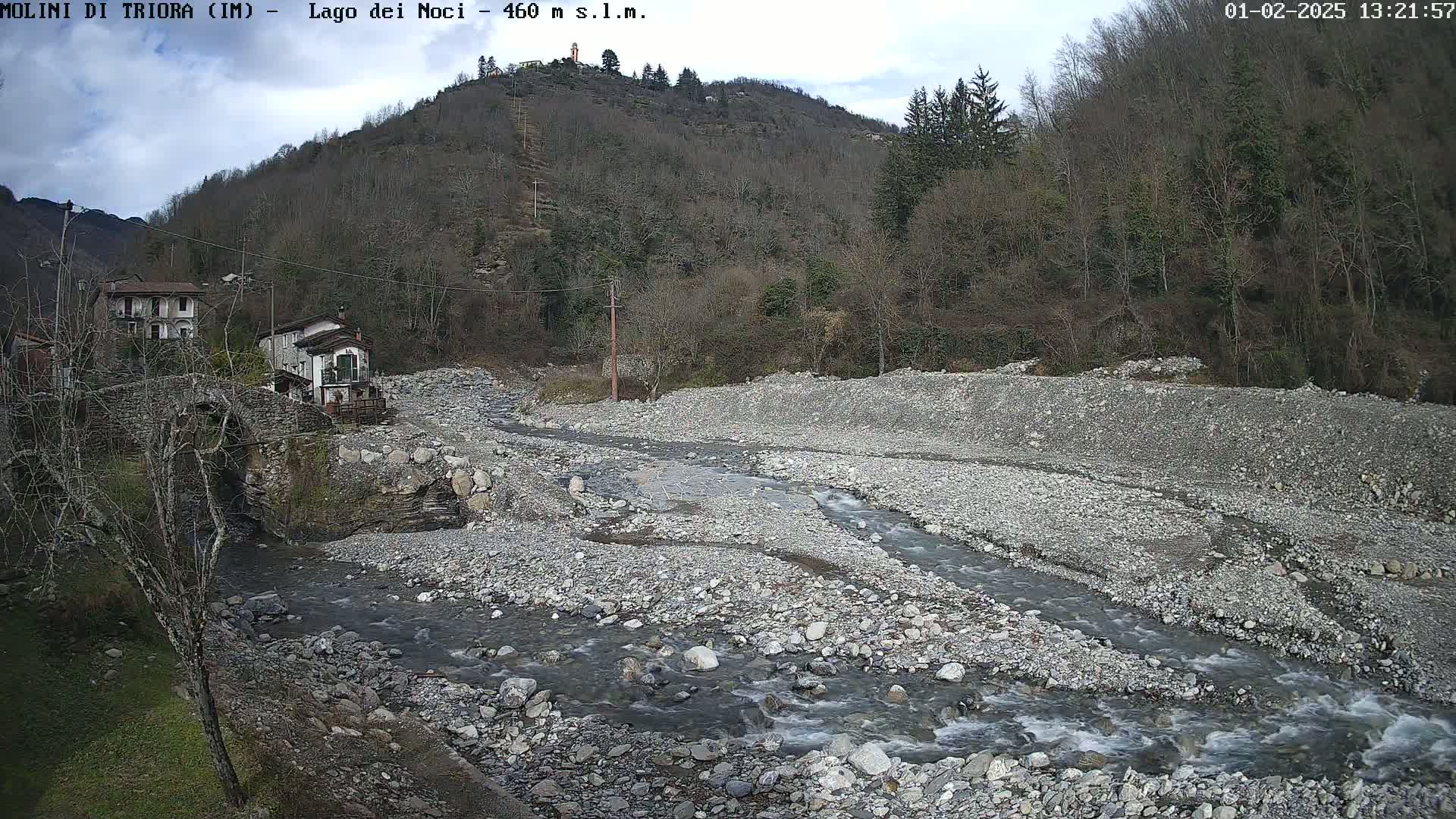 A rocky riverbed with a small stream flowing through it, nestled between buildings and a hillside of leafless trees under a mostly cloudy sky.