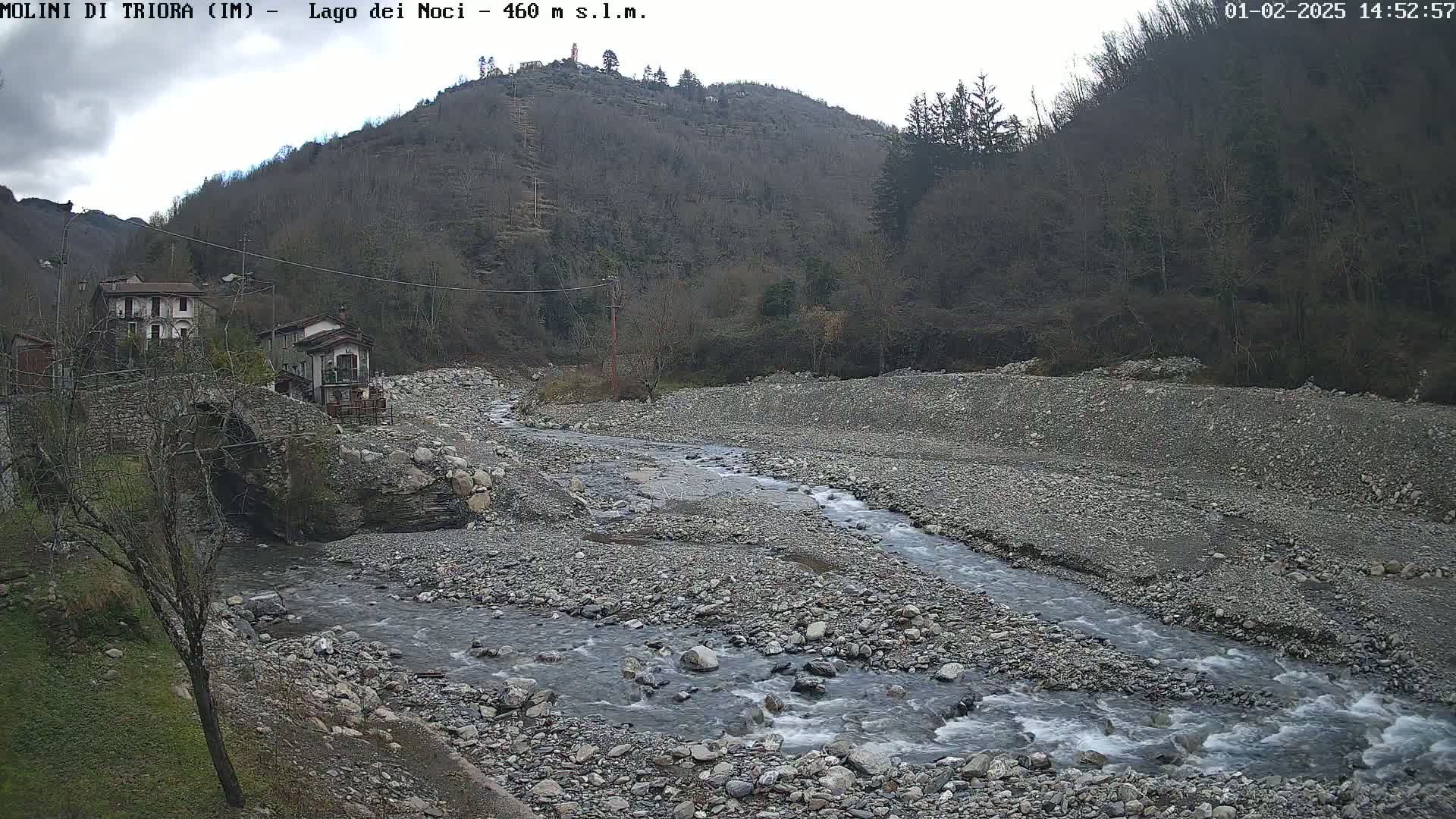 A rocky river flows through a valley under an overcast sky, passing under a stone bridge near a small village nestled against a hillside of leafless trees.