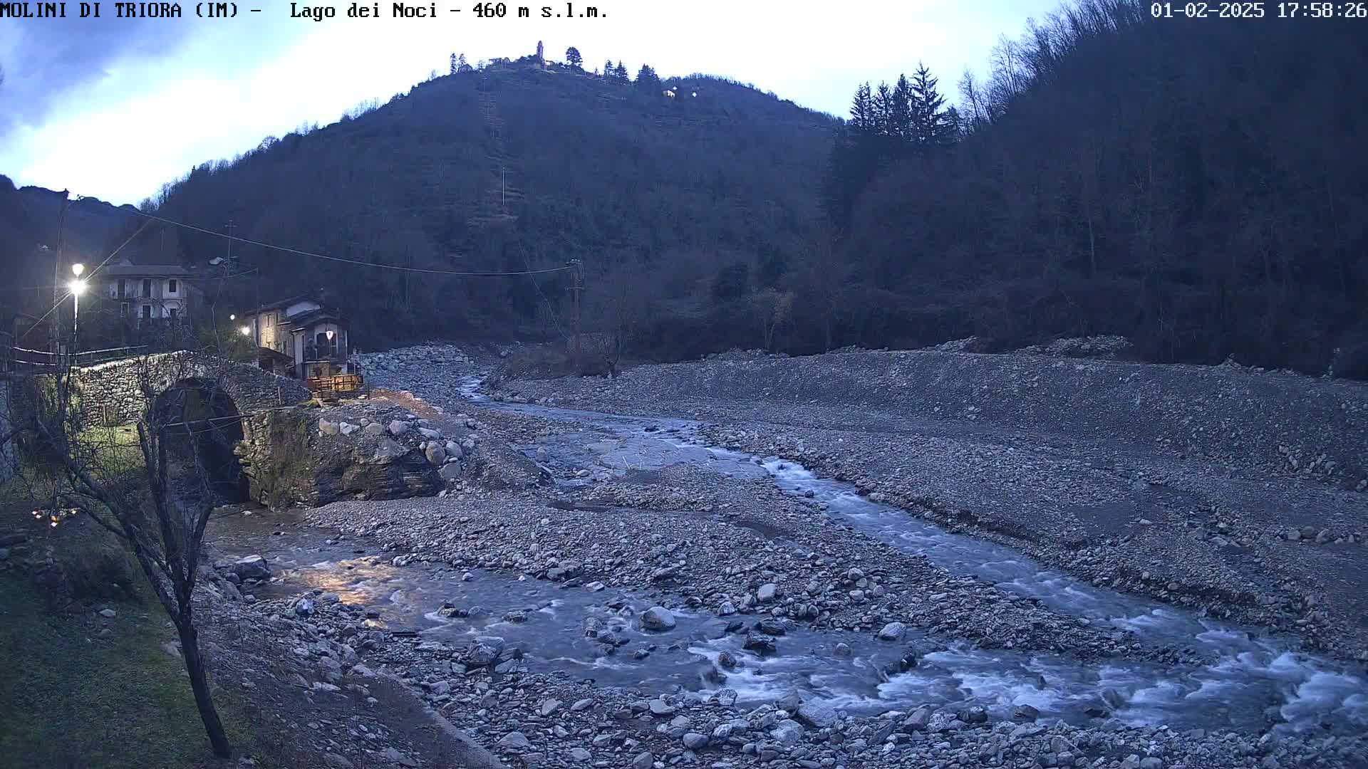 A rocky stream flows through a valley at twilight, passing under a stone arch bridge near buildings nestled against a hillside.