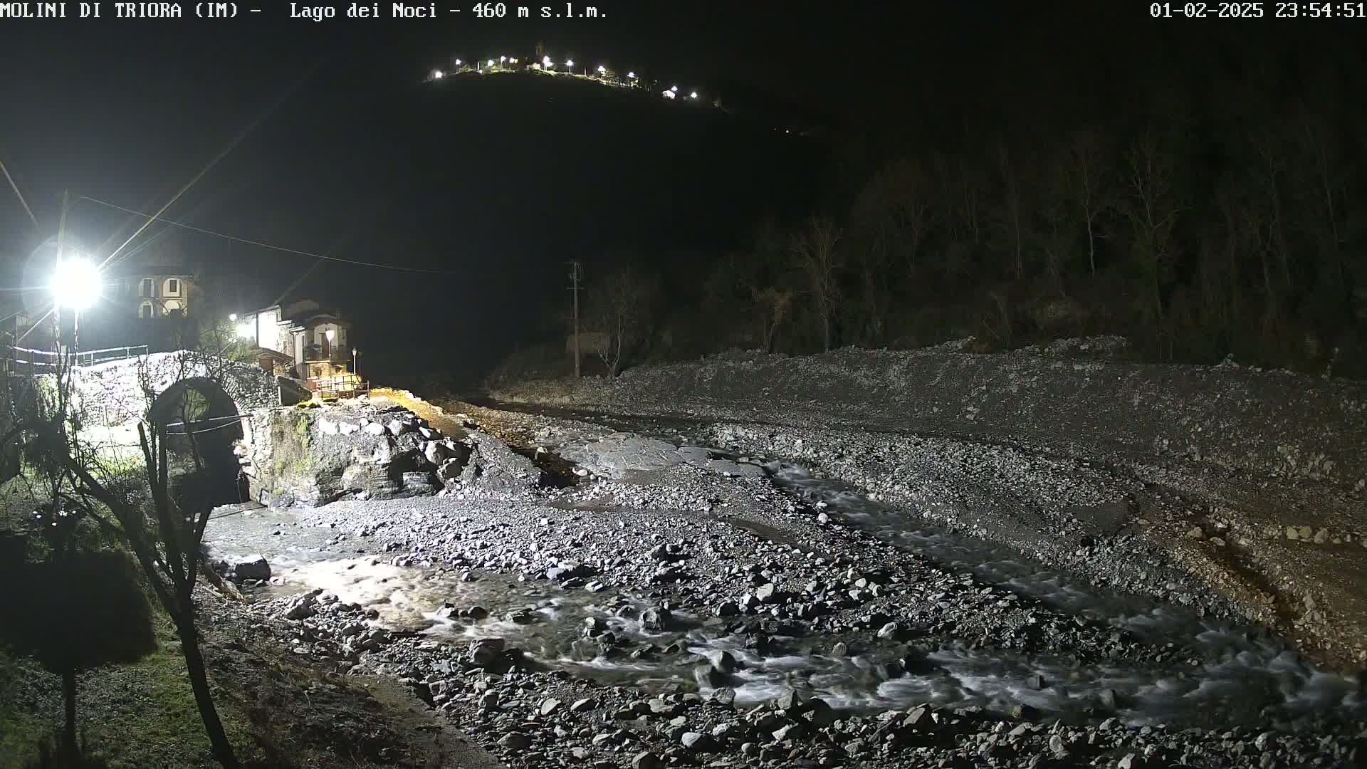A nighttime view of a stone bridge over a rocky stream, with a village visible in the background under artificial light.