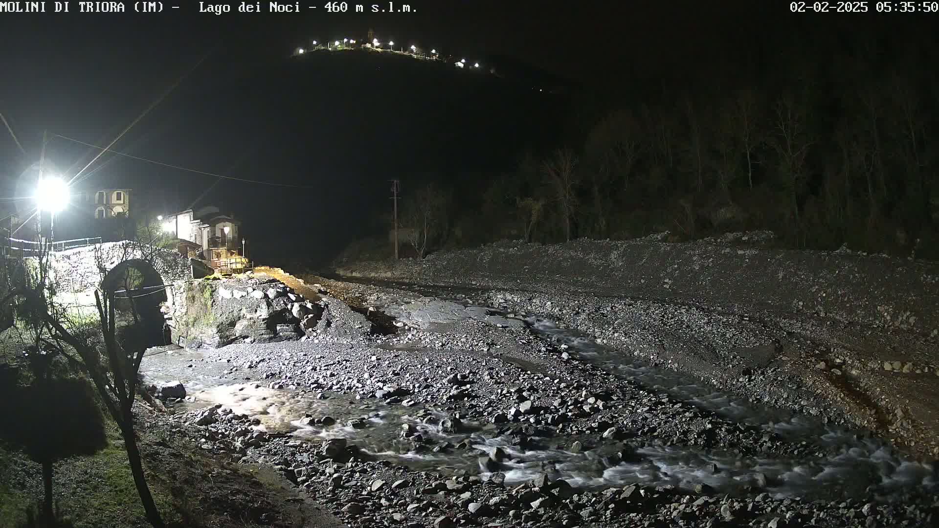 A nighttime view of a stone bridge over a rocky stream, with a village visible in the background under illuminated lights.