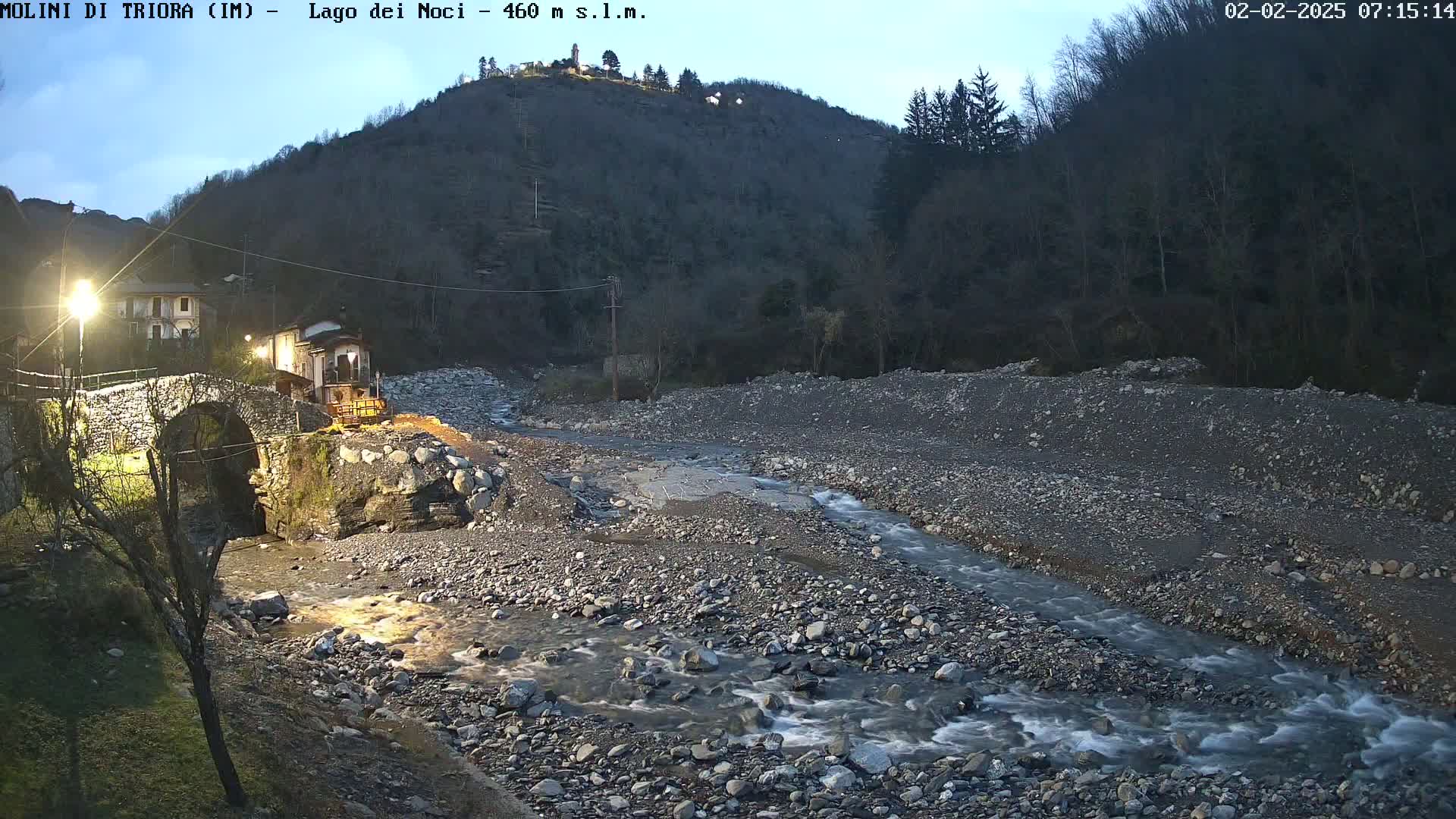 A stone bridge crosses a shallow, rocky river at dusk, under a dark, hilltop forest.