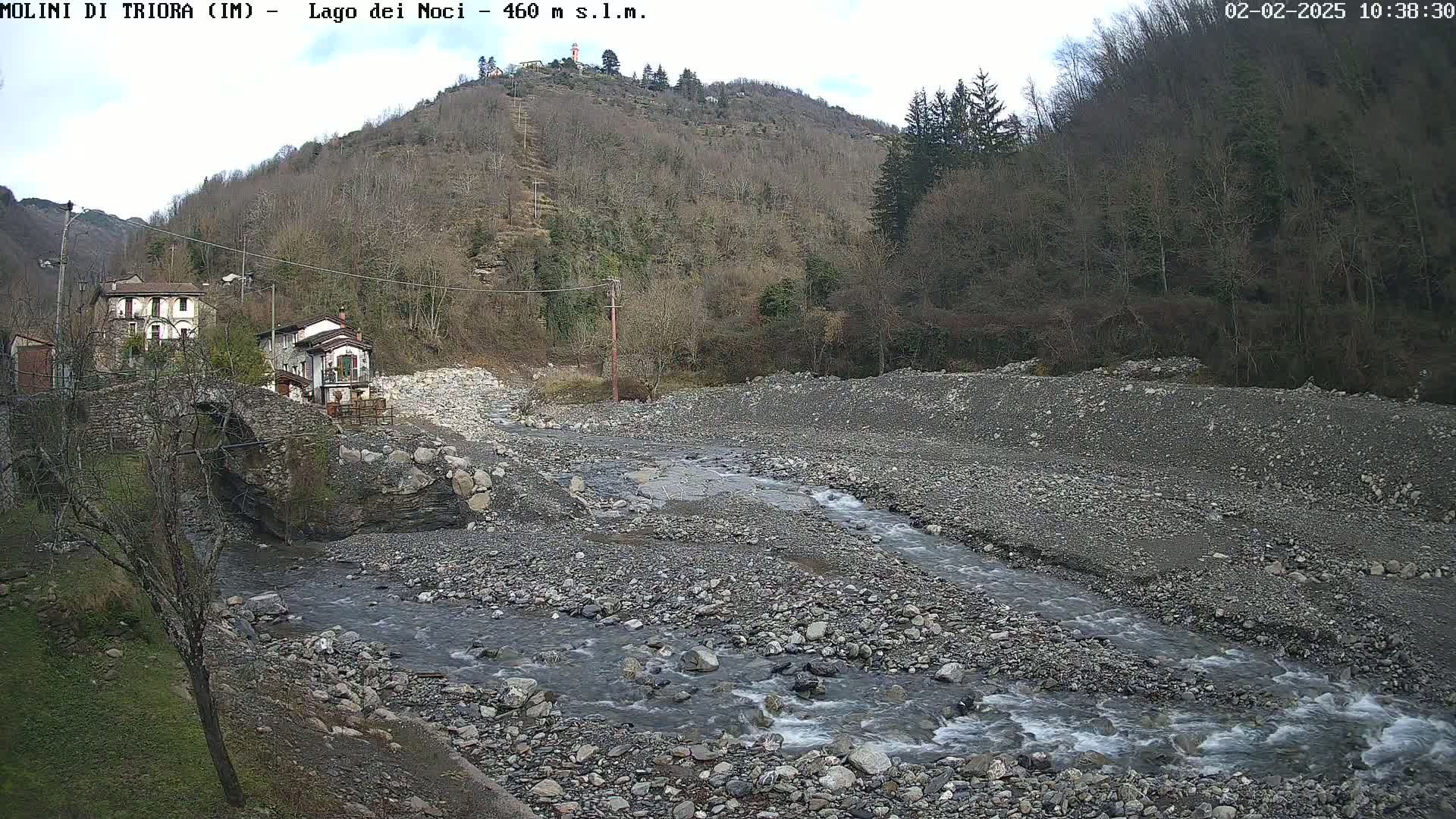 A rocky, shallow stream flows through a mostly barren riverbed, past a stone bridge and two buildings nestled against a hillside under a partly cloudy sky.