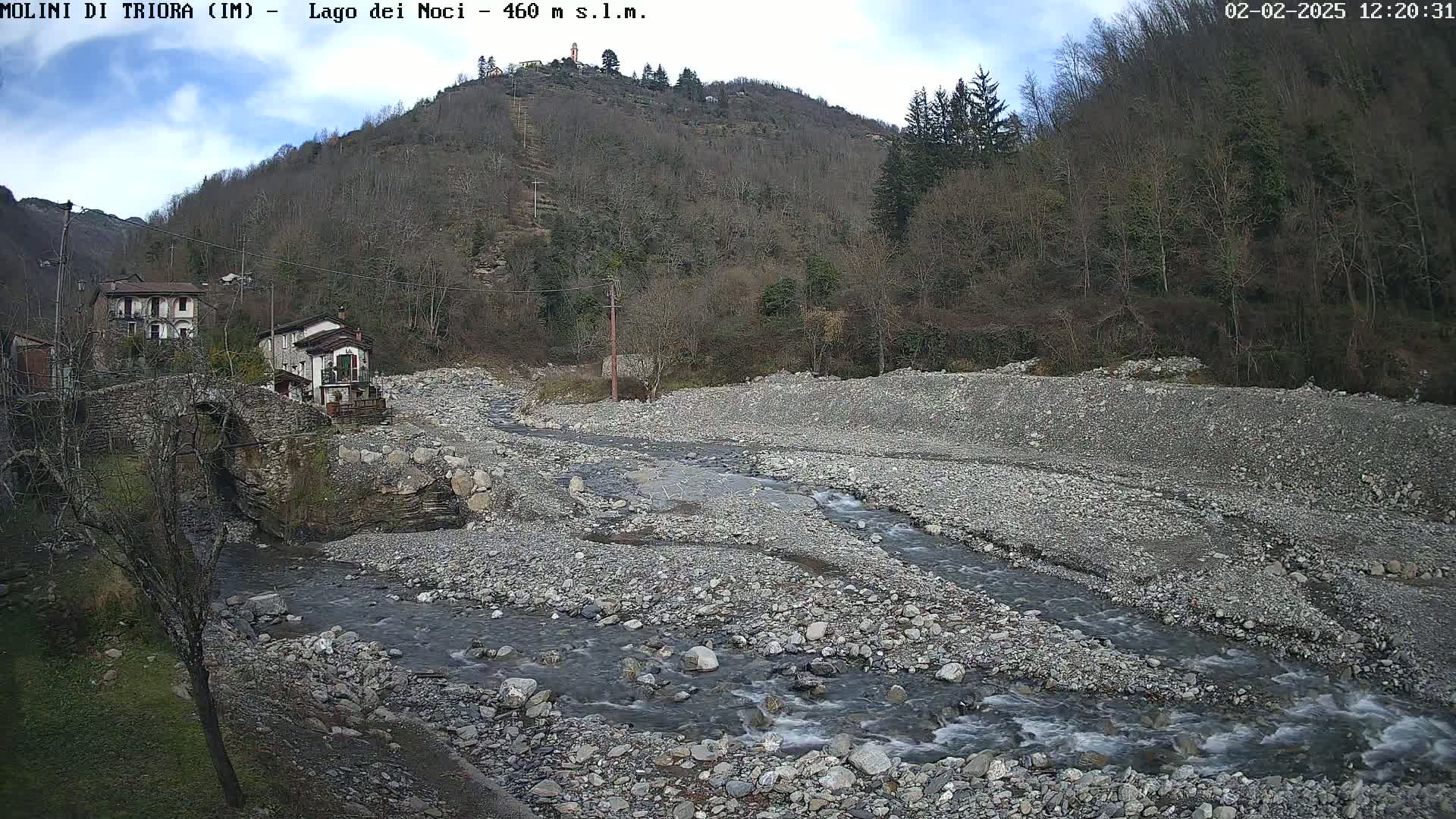 A rocky, shallow river flows through a mostly dry riverbed near a stone bridge and village nestled against a hillside under a partly cloudy sky.