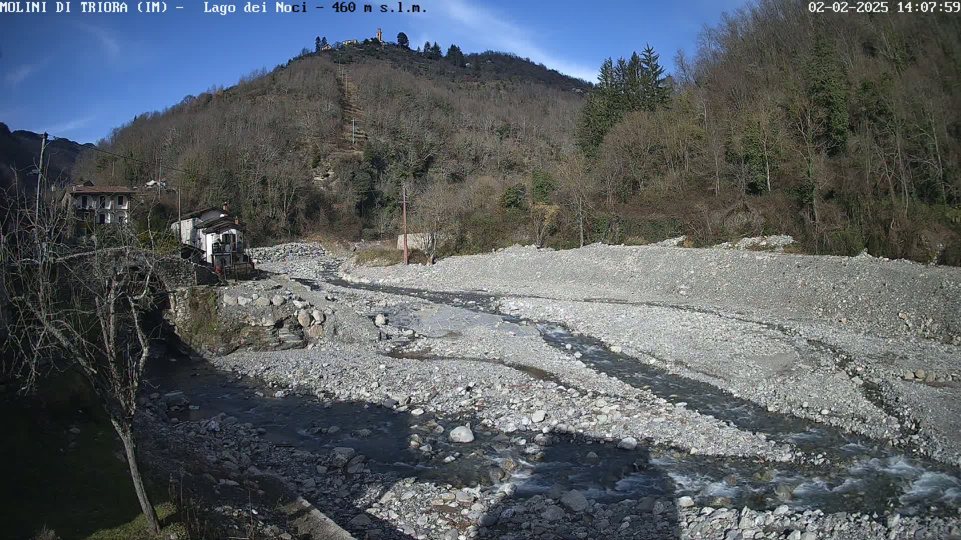 A rocky, mostly dry riverbed with a small stream flowing through it is situated below a hillside covered in leafless trees under a clear, sunny sky.