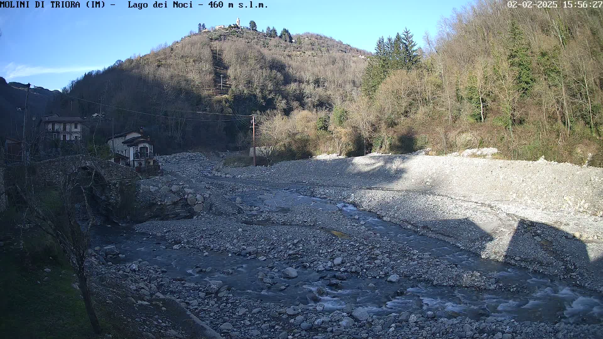 A rocky river flows through a valley under a clear blue sky, past a stone bridge and several buildings nestled against a hillside covered in leafless trees.