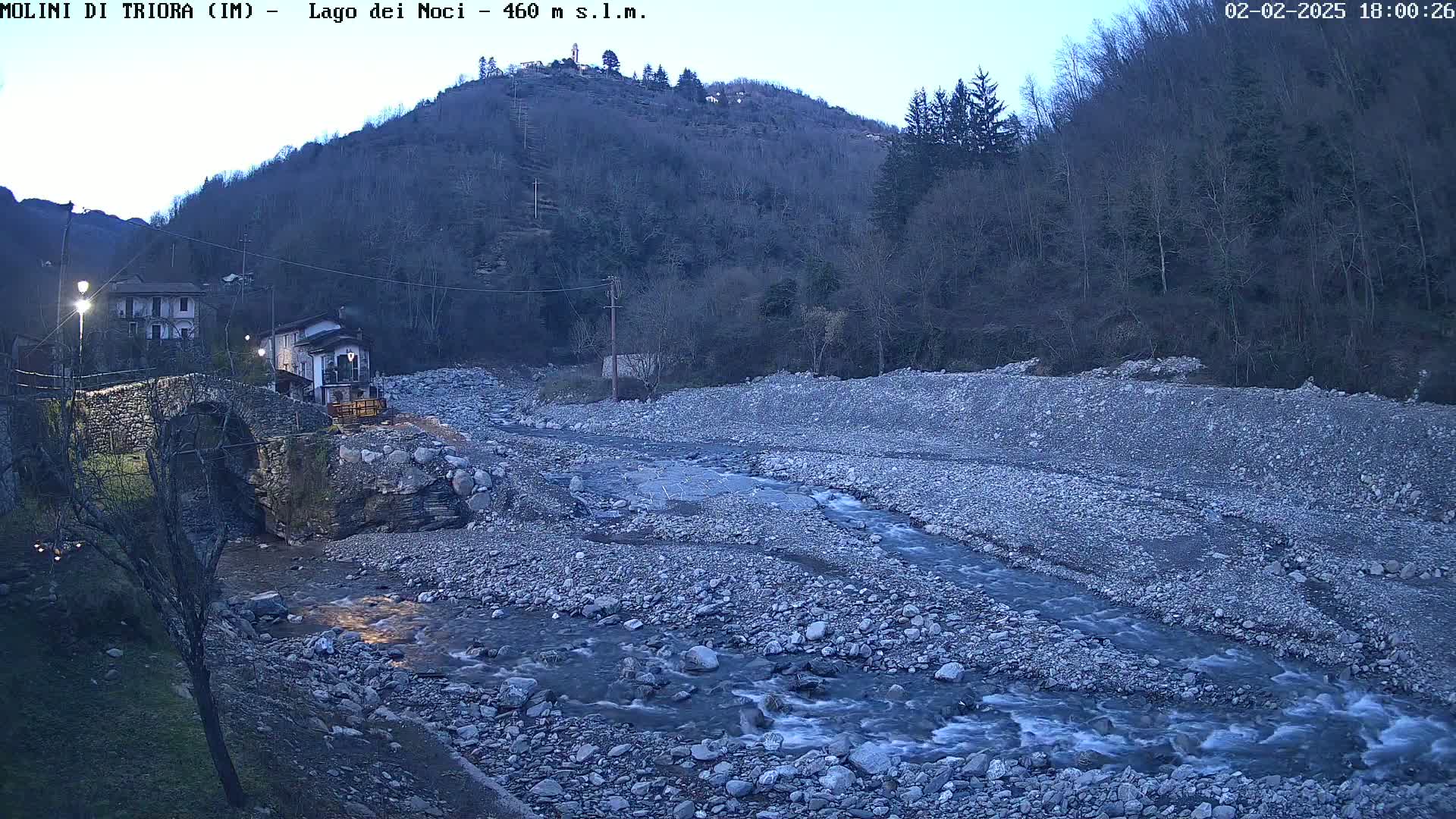 A rocky riverbed with a flowing stream is seen beneath a stone bridge and buildings nestled in a hilltop forest at dusk.