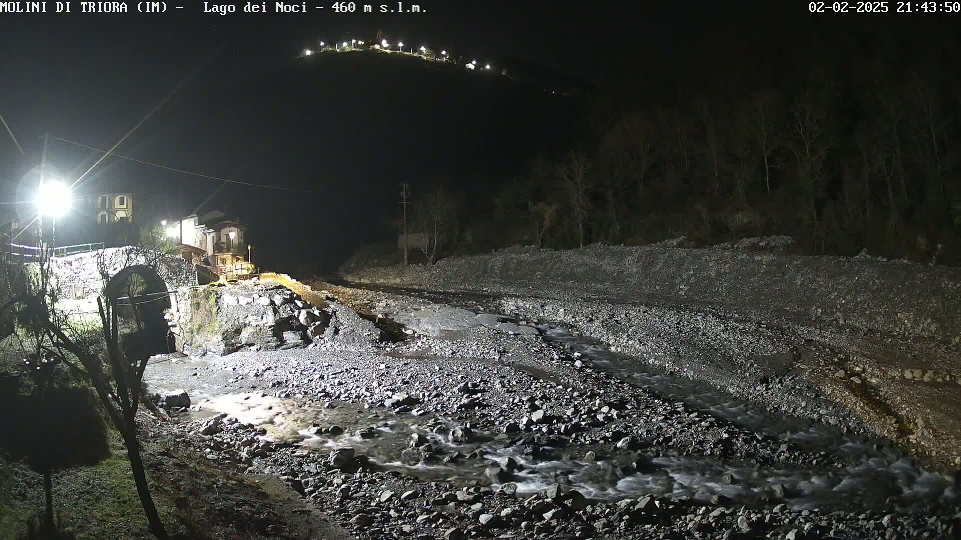 A nighttime view of a rocky riverbed with a small stream flowing through it, a stone bridge, and buildings illuminated by artificial light under a dark sky.