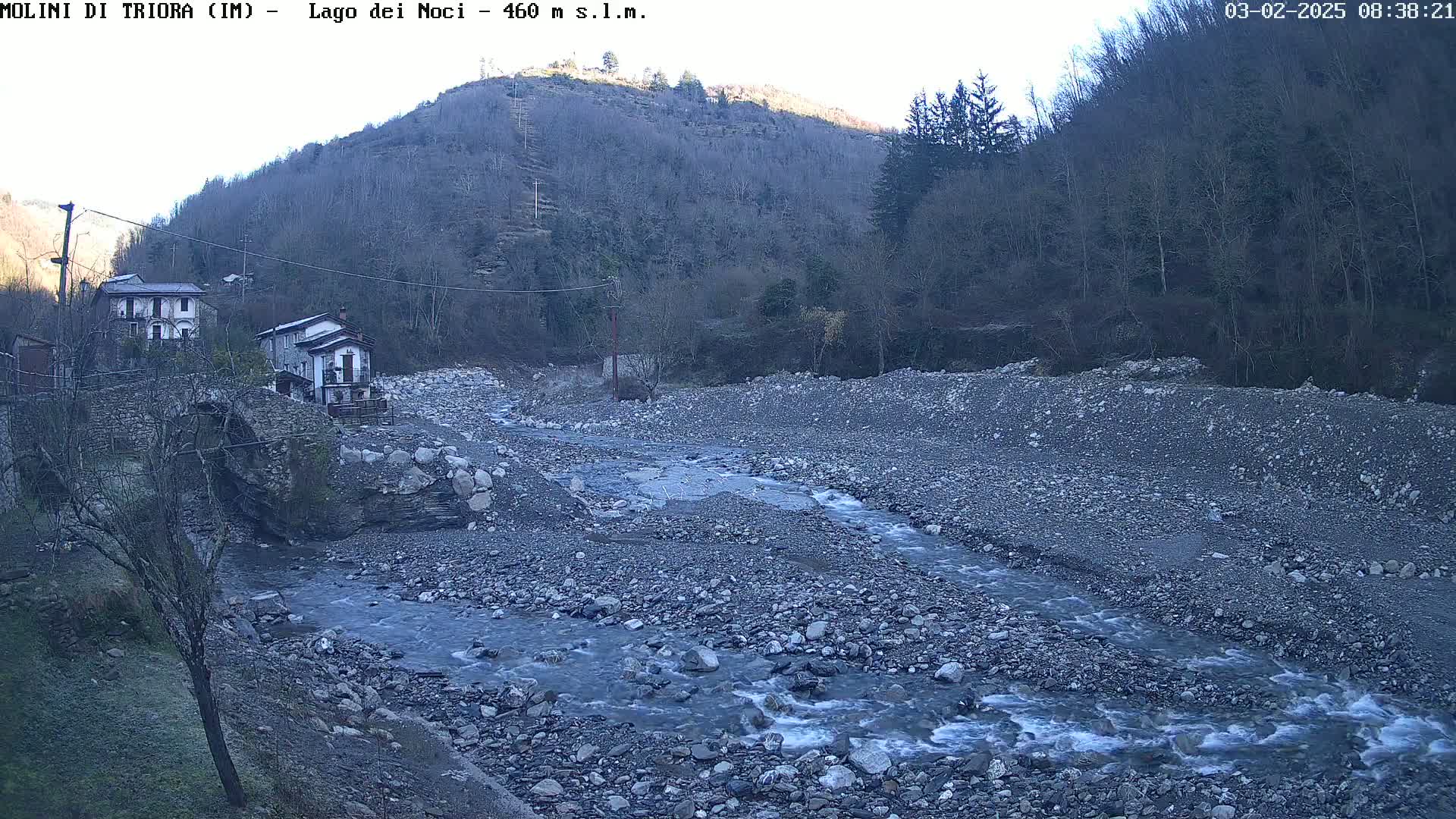 A rocky river flows past two houses nestled at the base of a mountain under a clear, bright sky.