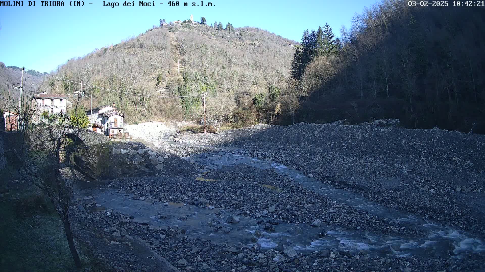 A rocky river flows through a valley under a clear, sunny sky, past a small village nestled at the base of a hillside covered in leafless trees.