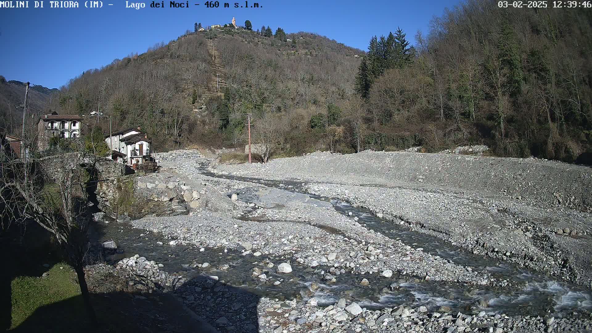A rocky, dry riverbed with a small stream running through it, under a clear blue sky, is situated between buildings and a hillside covered in leafless trees.