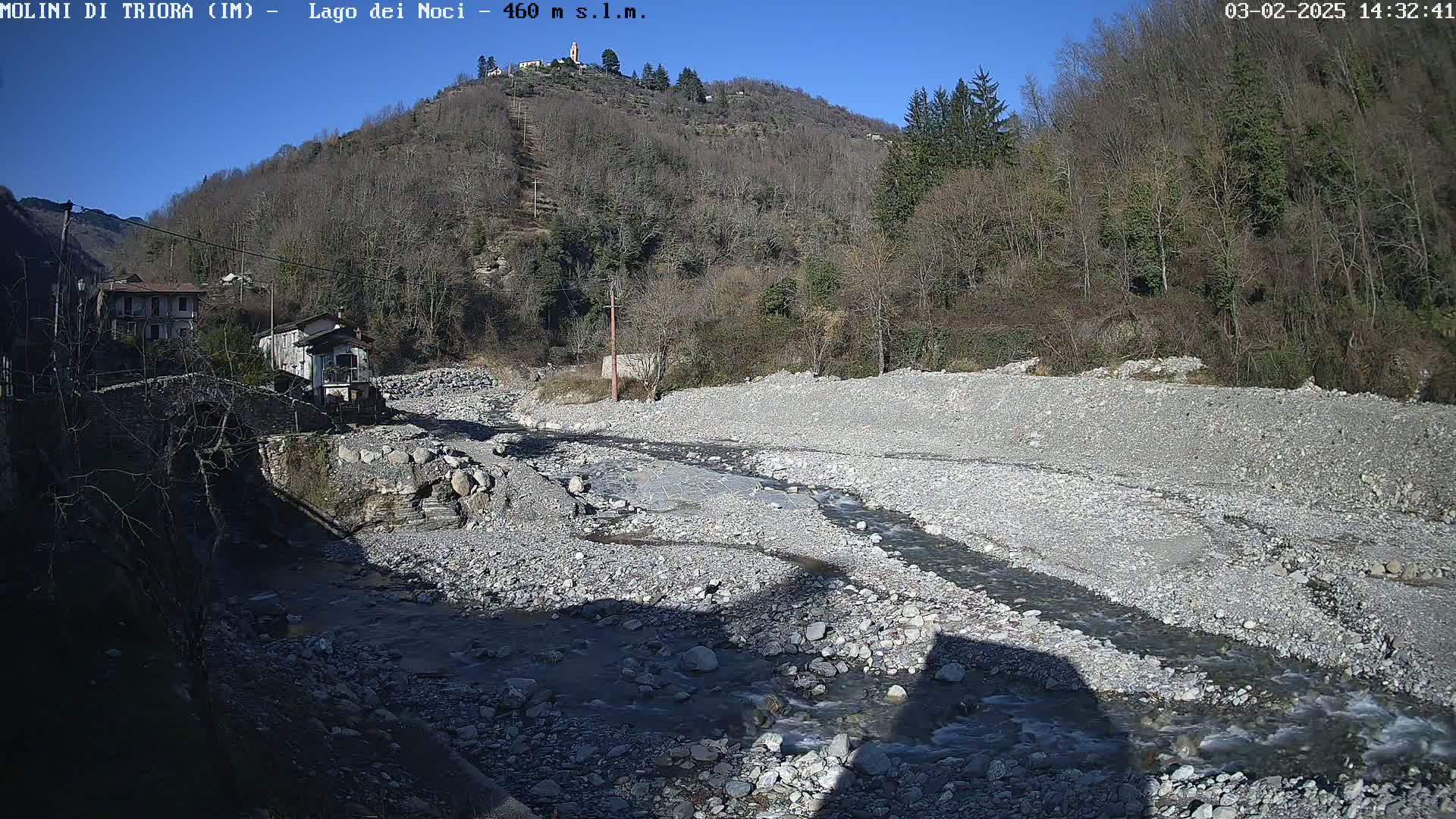 A mostly dry, rocky riverbed with a small stream meandering through it, nestled between buildings and a hillside covered in leafless trees under a clear, sunny sky.