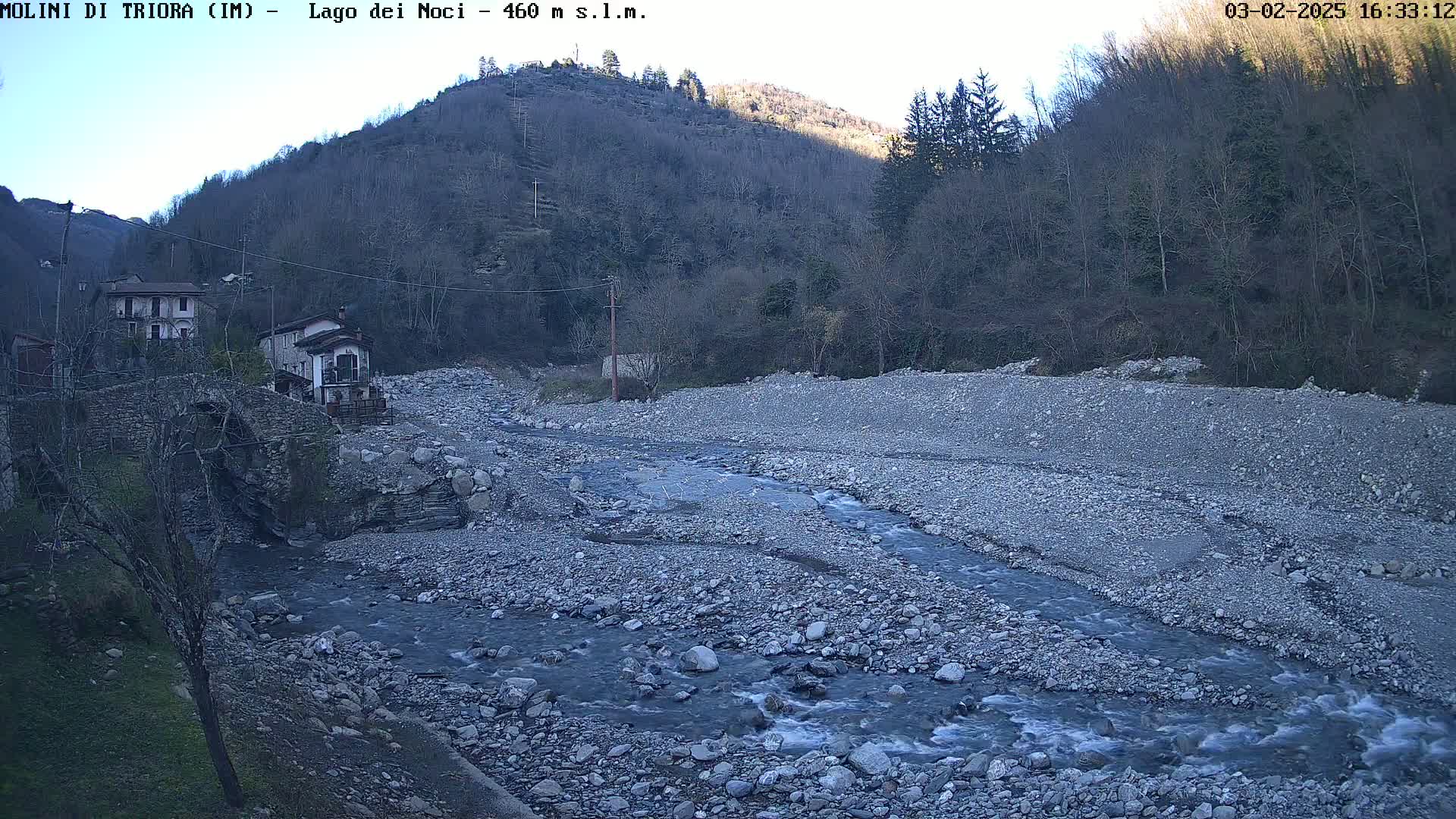 A rocky riverbed with a slow-moving stream flows past a stone bridge and houses nestled against a hillside under a clear sky.