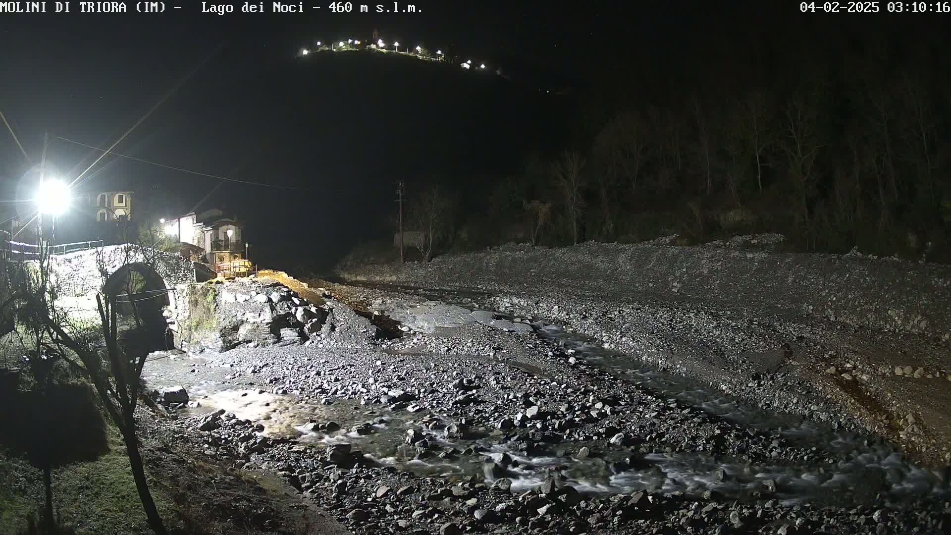 A nighttime view of a rocky riverbed with a stone bridge and buildings under artificial light, with a hill in the background.