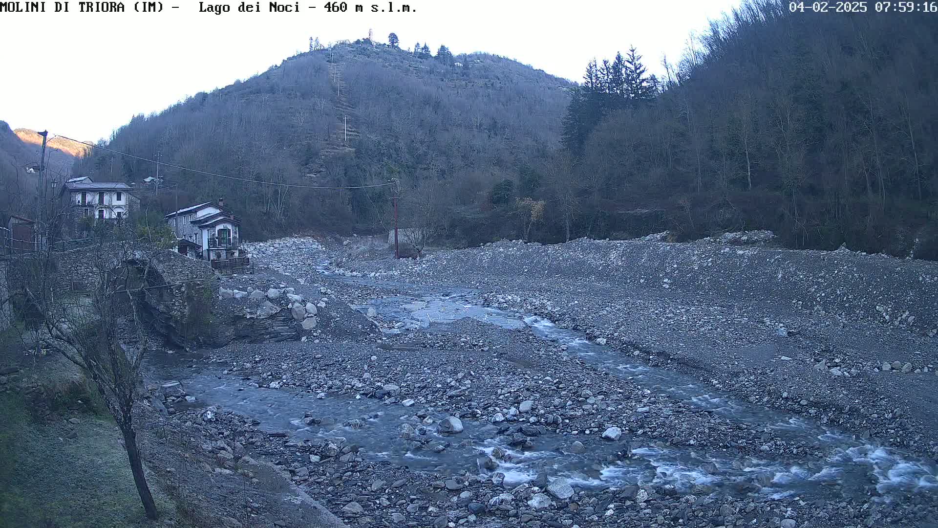 A rocky, shallow river flows past two houses and a stone bridge, nestled in a valley with leafless trees under a clear, dawn sky.