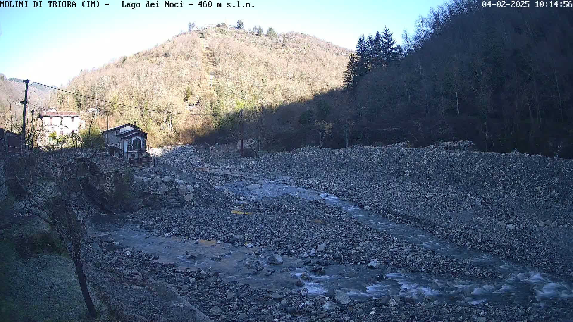 A rocky riverbed with a shallow stream flows through a valley under a clear, sunny sky, past a stone bridge and buildings nestled against a hillside covered in leafless trees.