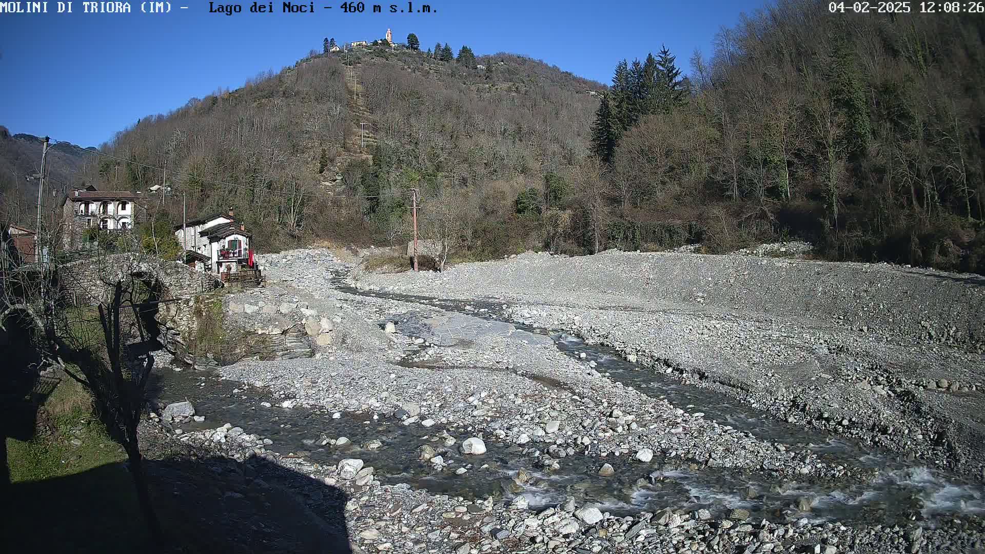 A rocky riverbed with a shallow, meandering stream flows past a stone bridge and several buildings nestled against a hillside under a clear, sunny sky.