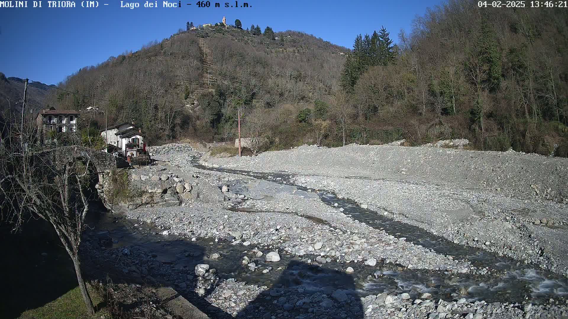 A rocky, dry riverbed with a small stream flowing through it, nestled in a valley between bare winter trees under a clear blue sky.