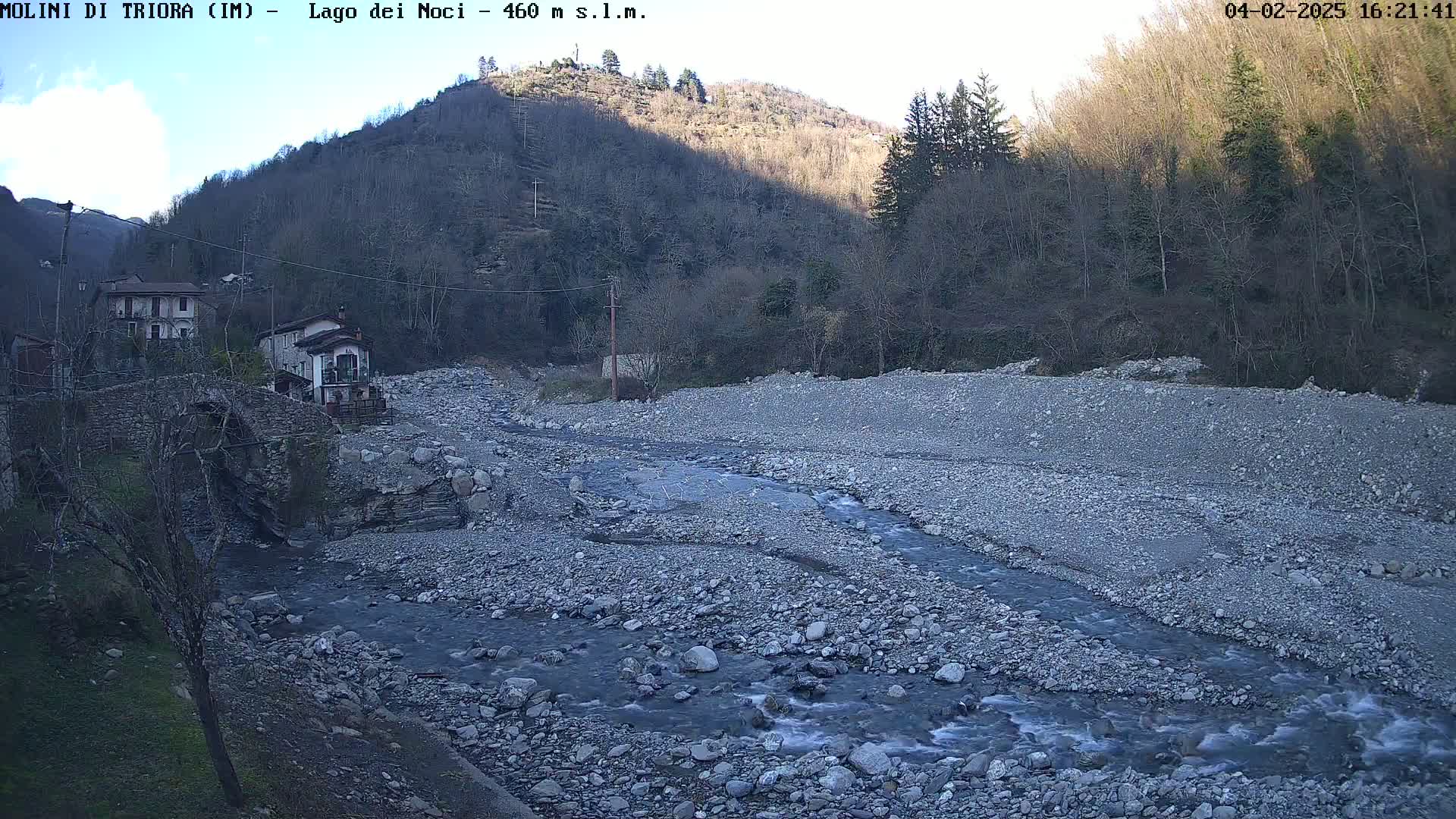 A rocky riverbed with a slow-moving stream flows past a stone bridge and small village nestled in a valley under a partly cloudy sky.