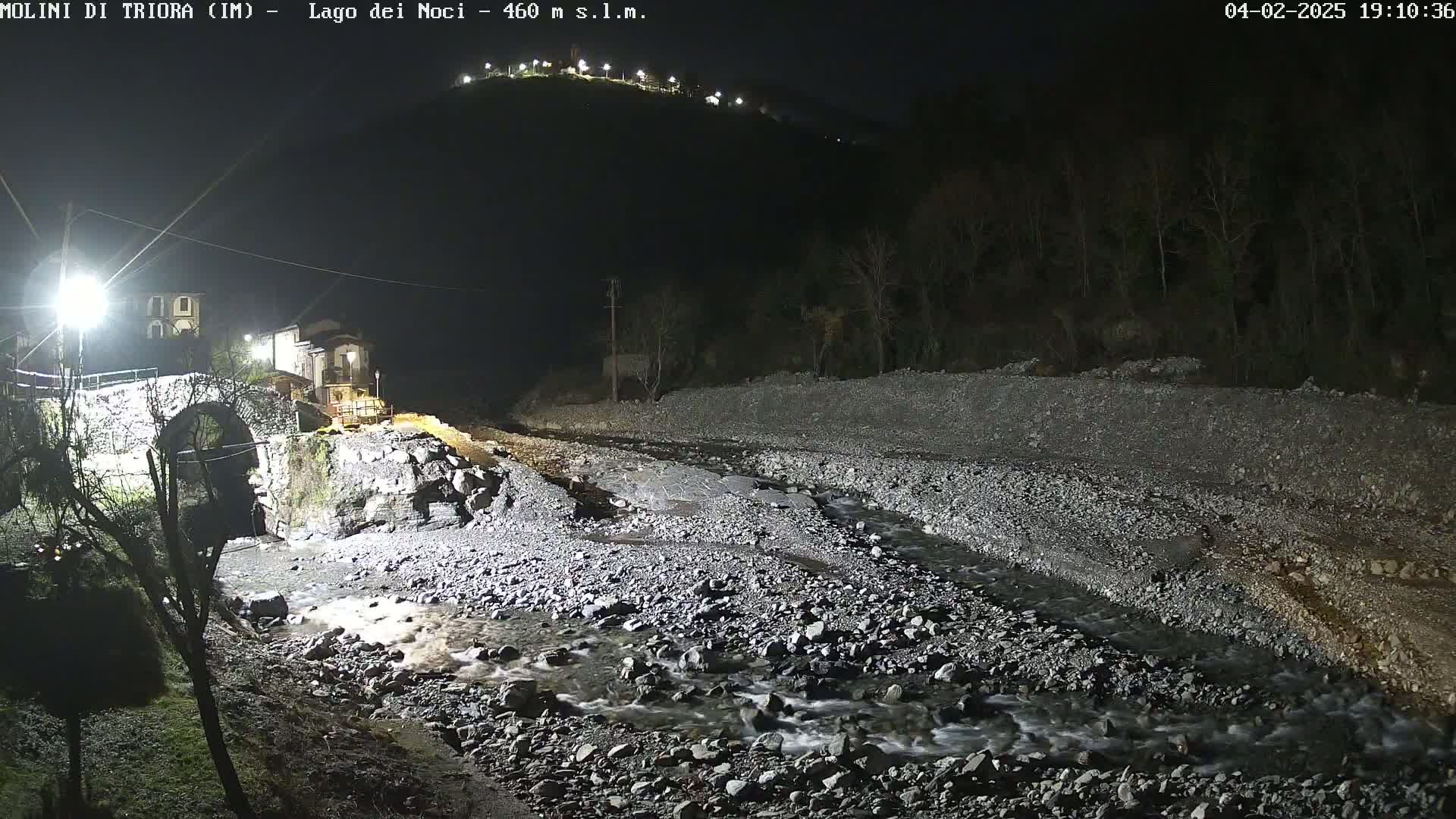 A rocky stream flows under a stone arch bridge at night, illuminated by artificial lights, with a hill in the background.