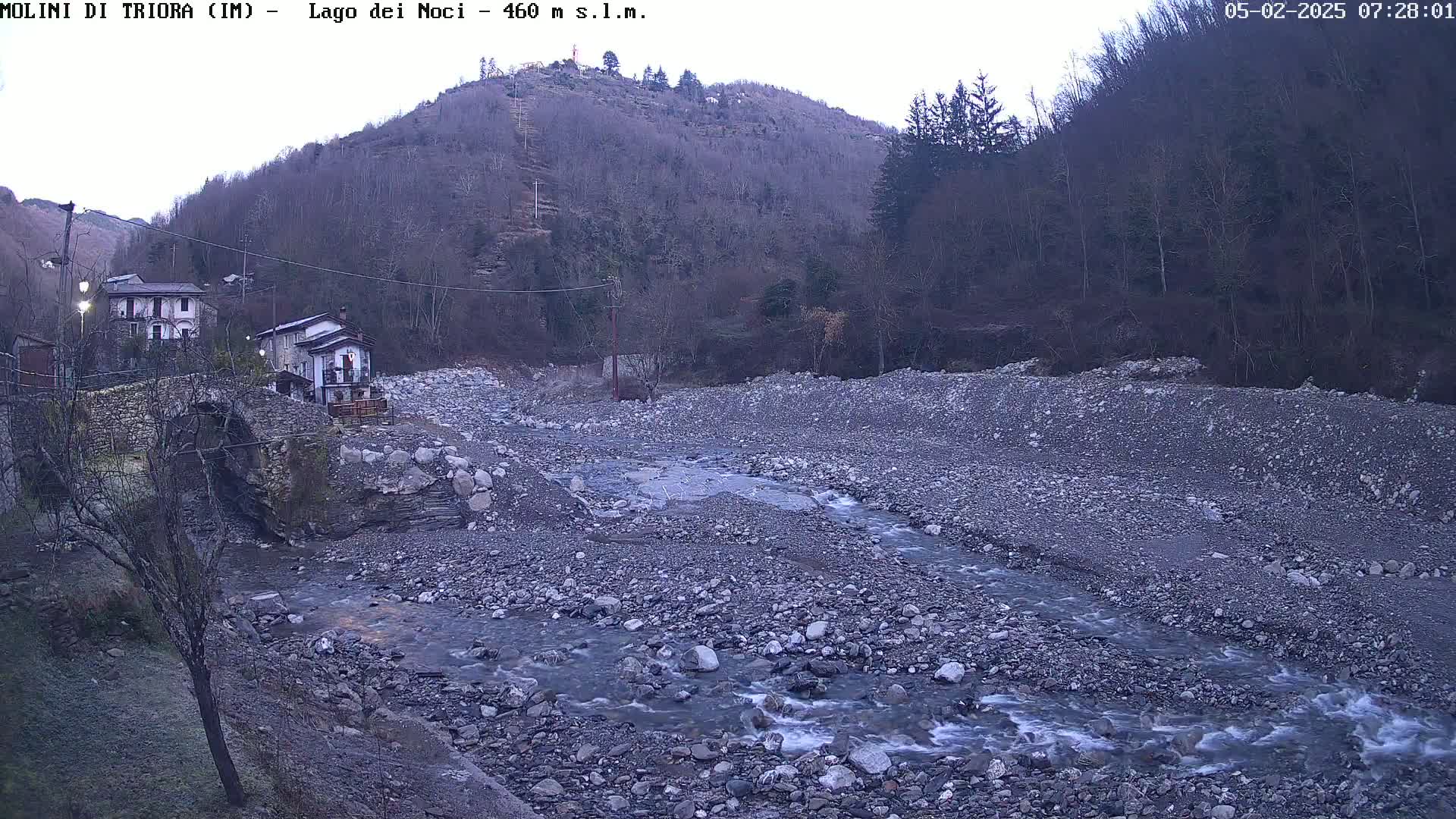 A rocky, shallow river flows through a valley under an overcast sky, passing by a stone bridge and buildings nestled against a hillside covered in leafless trees.