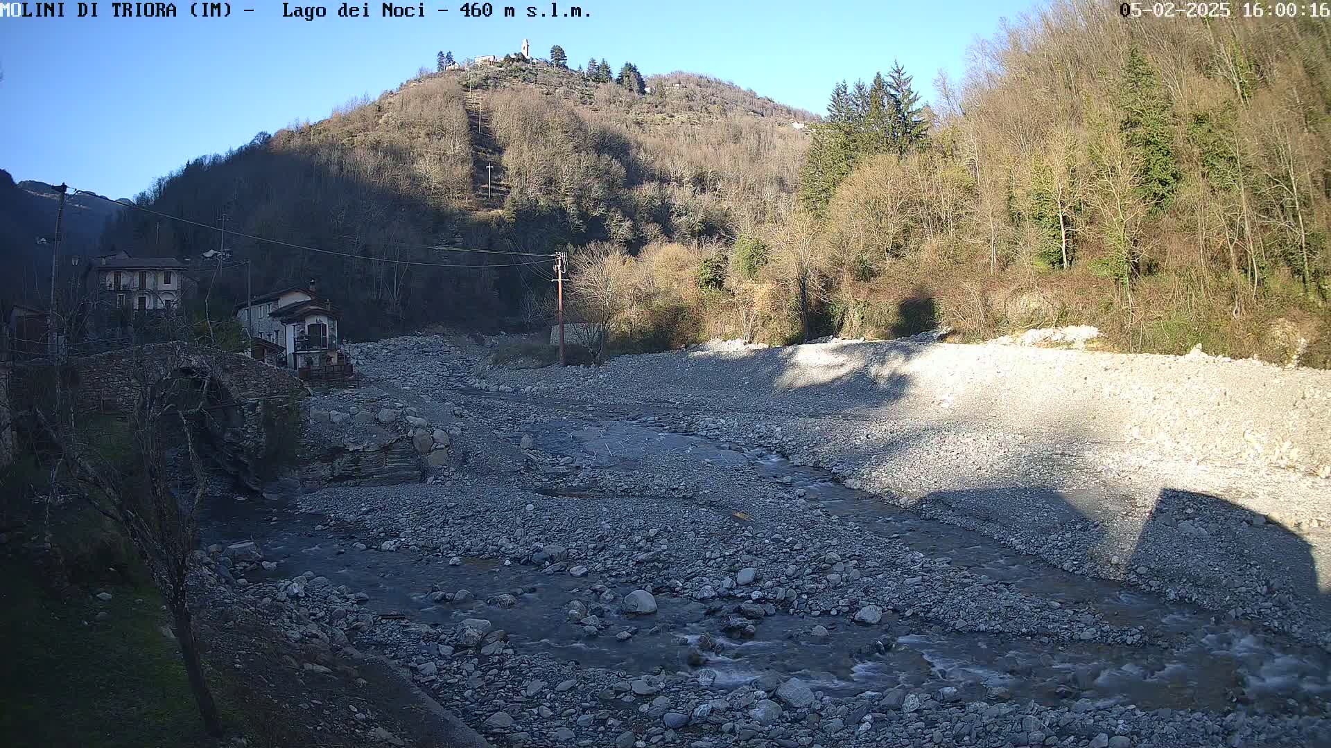 A rocky, shallow river flows through a dry riverbed, past buildings and a stone bridge, nestled in a valley under a clear, sunny sky.