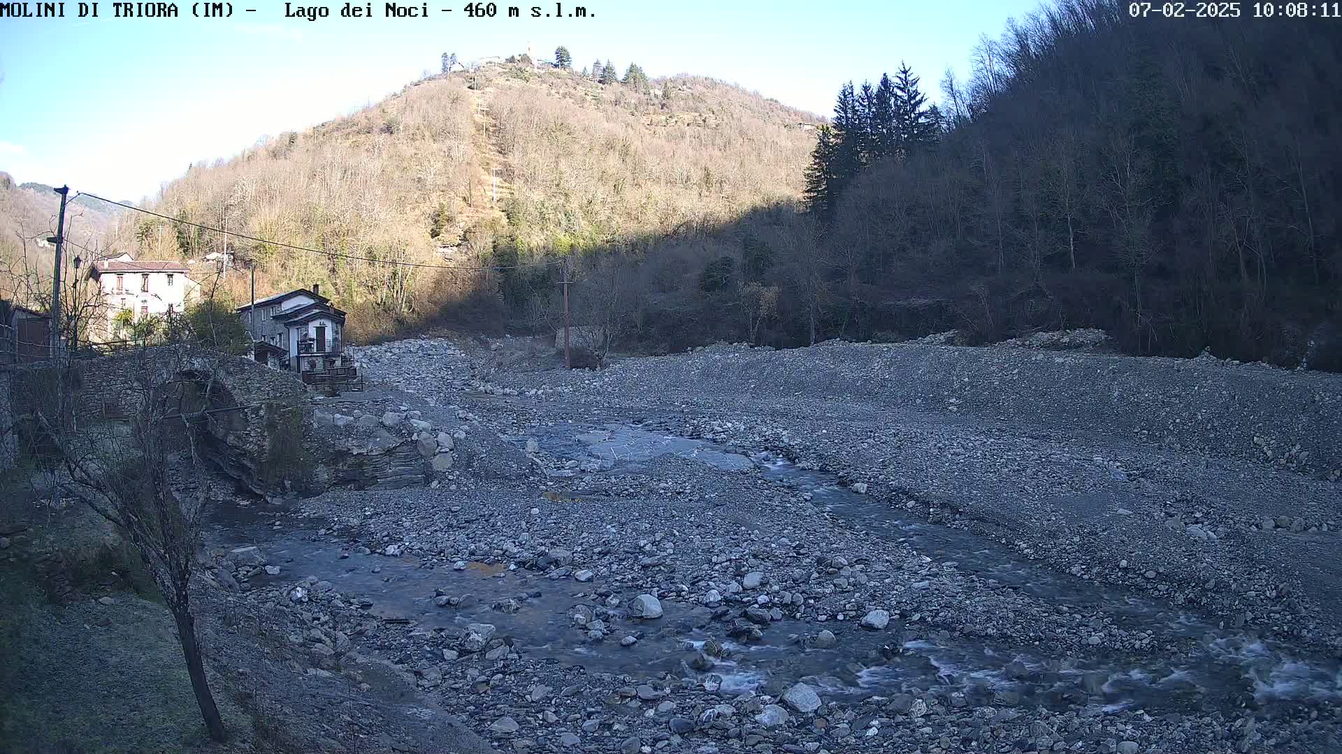 A rocky, shallow river flows through a mostly dry riverbed, past a stone bridge and buildings nestled at the base of a leafless, sunlit hillside under a clear sky.