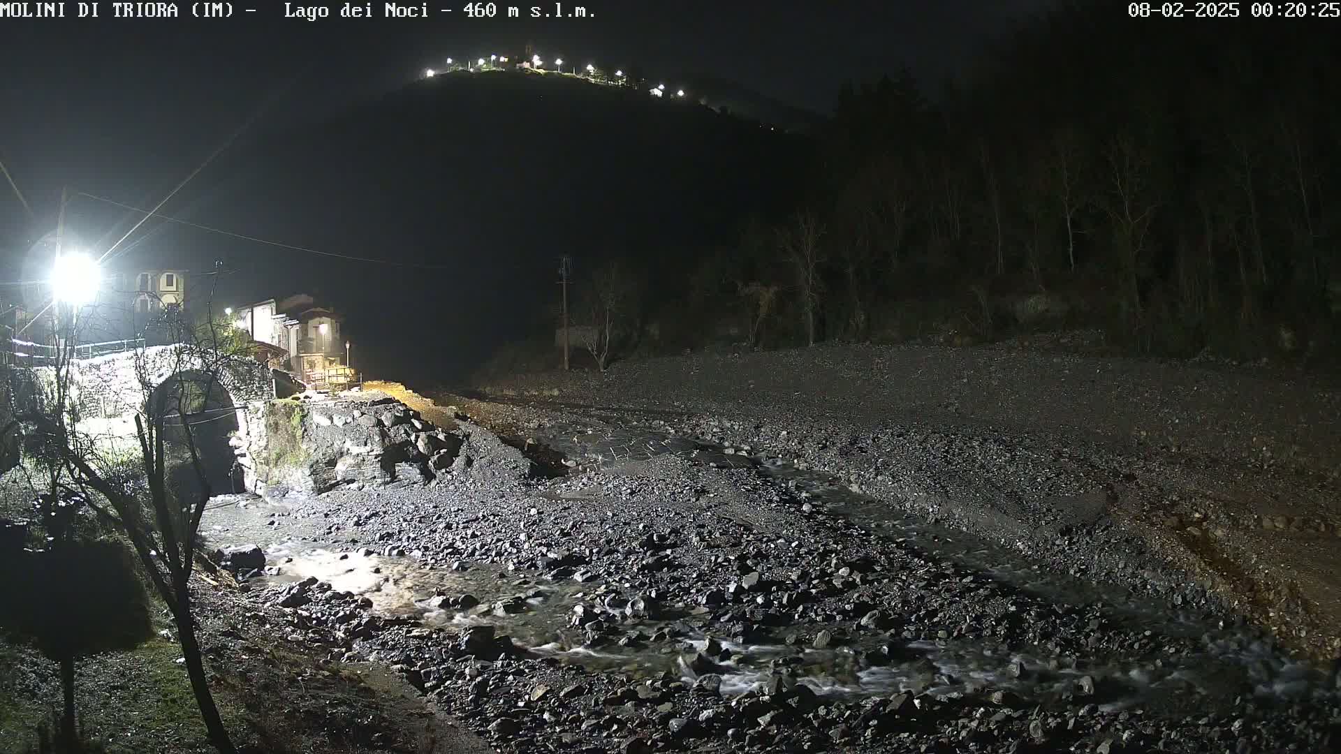 A nighttime view of a rocky stream flowing under a stone arch bridge near a small village, illuminated by artificial lights.