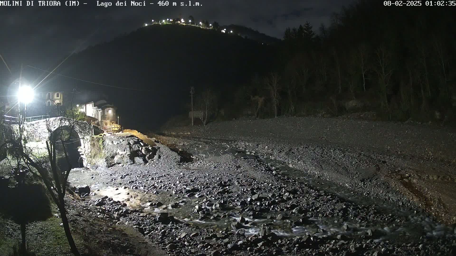 A nighttime view of a rocky stream flowing under a stone bridge near a small village, nestled against a dark hillside.