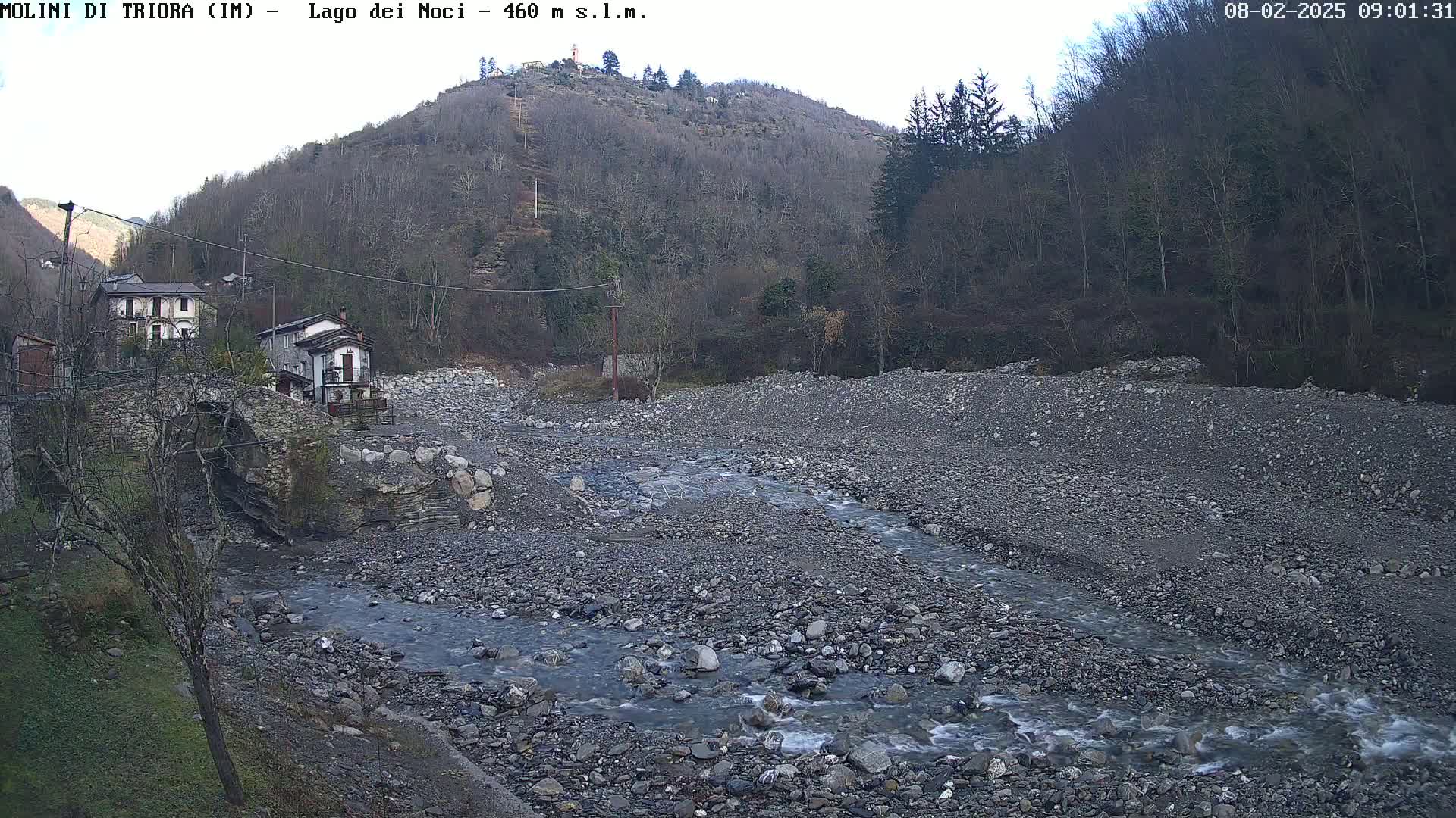 A rocky riverbed with a shallow stream flows past a stone bridge and houses nestled against a hillside under a partly cloudy sky.