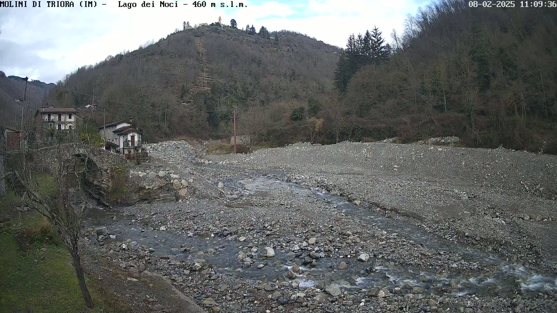 A rocky, shallow river flows through a valley under a partly cloudy sky, past a stone bridge and small village nestled against a hillside covered in leafless trees.