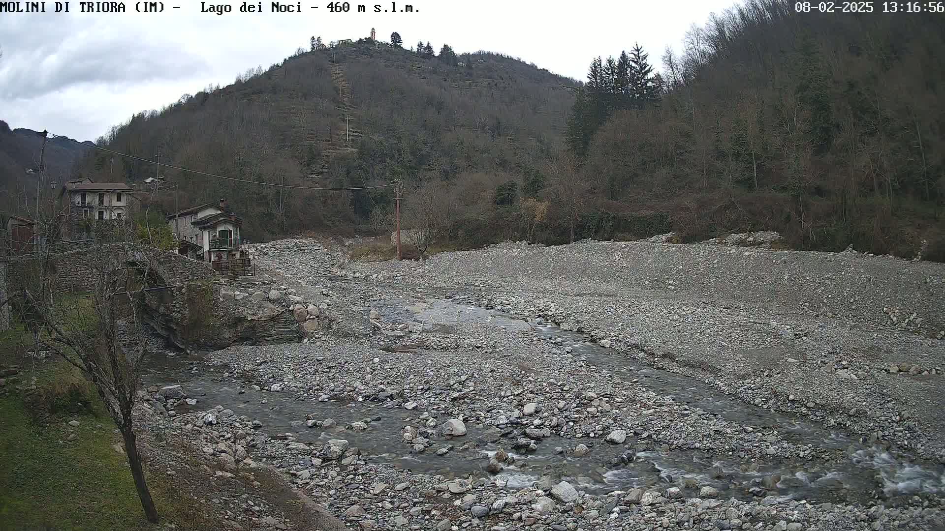 A rocky riverbed with a shallow stream flows past a stone bridge and buildings nestled against a hillside under an overcast sky.
