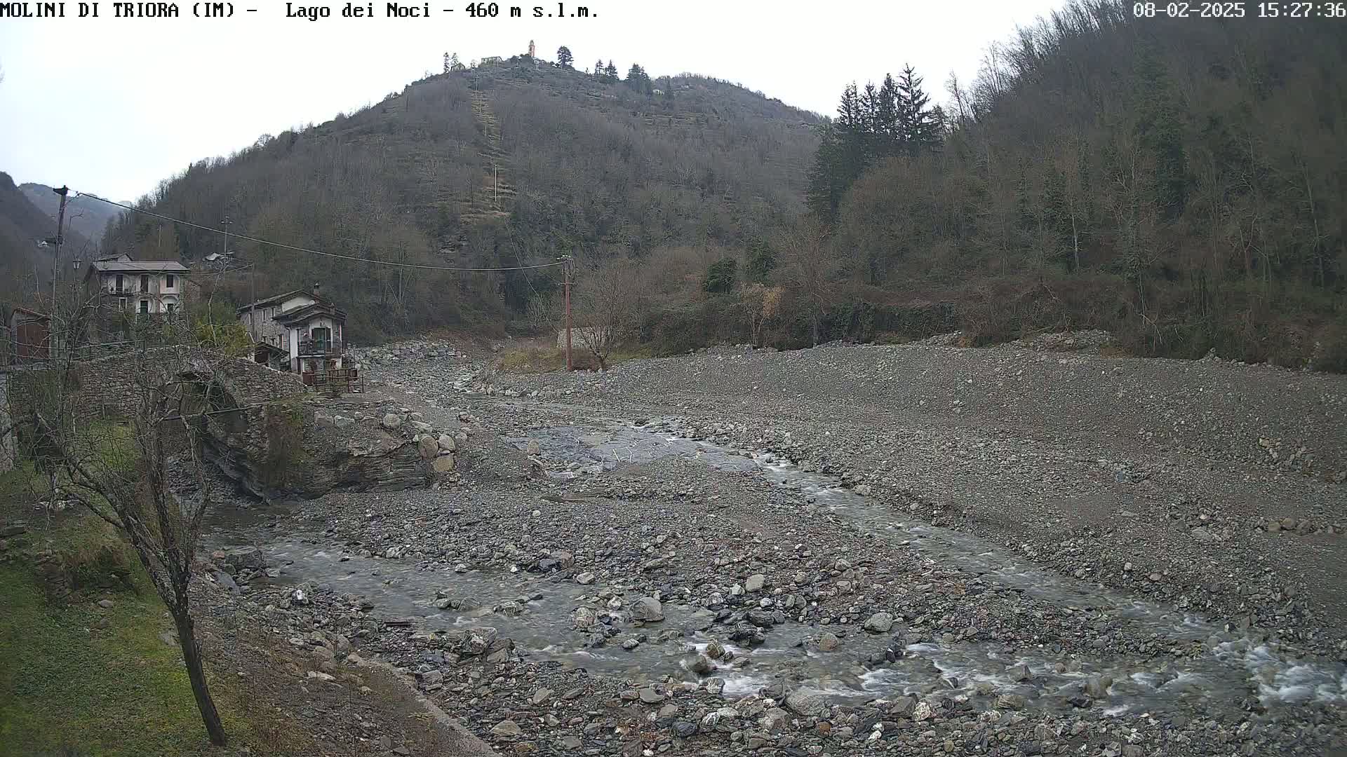 A rocky, shallow river flows through a dry riverbed, past a small village nestled at the base of a wooded hillside under an overcast sky.