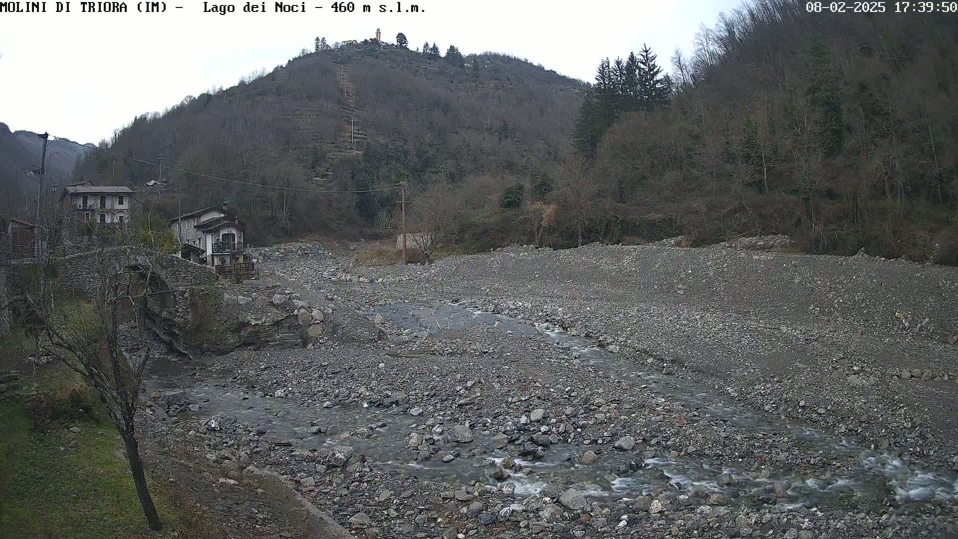 A rocky, shallow stream flows through a sparsely vegetated valley under an overcast sky, past a stone bridge and several buildings nestled against a hillside.