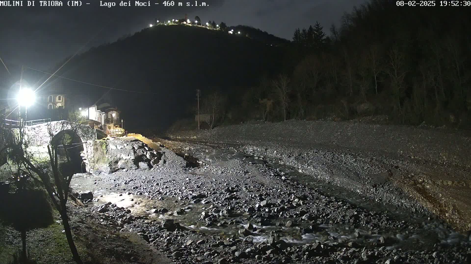 A nighttime view of a stone bridge over a shallow, rocky stream, with buildings and a hill visible in the background under a dark sky.