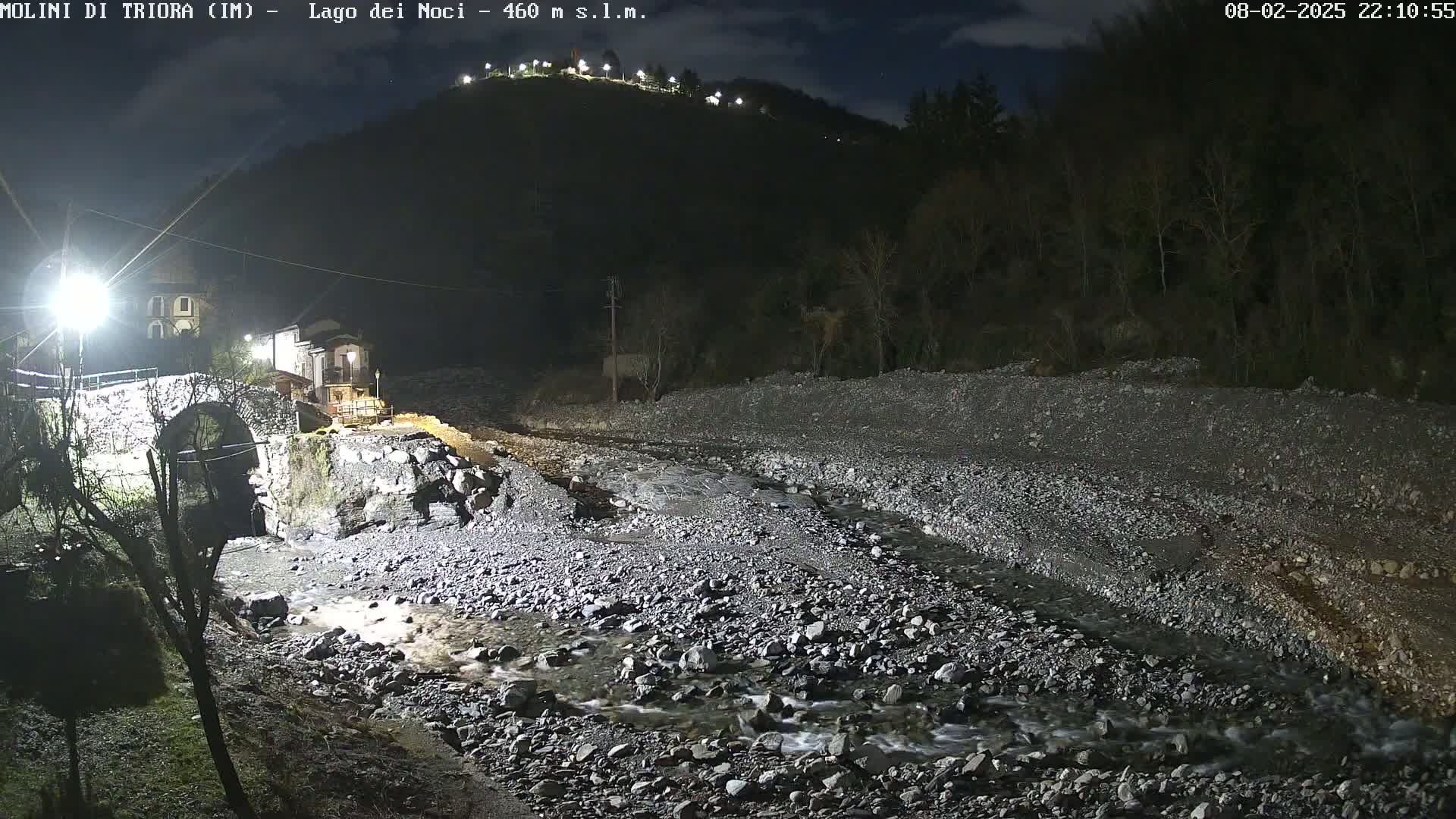 A nighttime view of a rocky riverbed with a stone bridge and buildings near a hill under a partly cloudy sky.