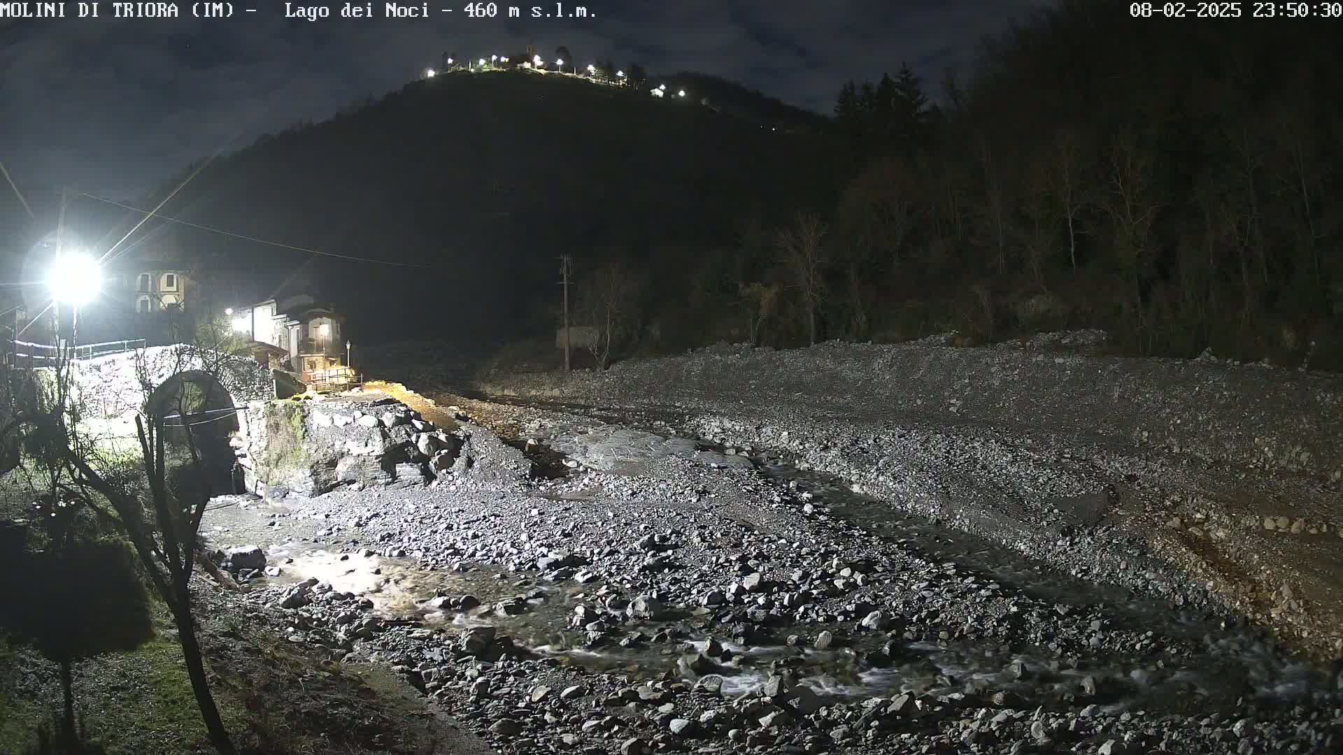 A stone bridge and buildings are illuminated at night next to a shallow, rocky stream flowing under a dark, tree-covered hill.