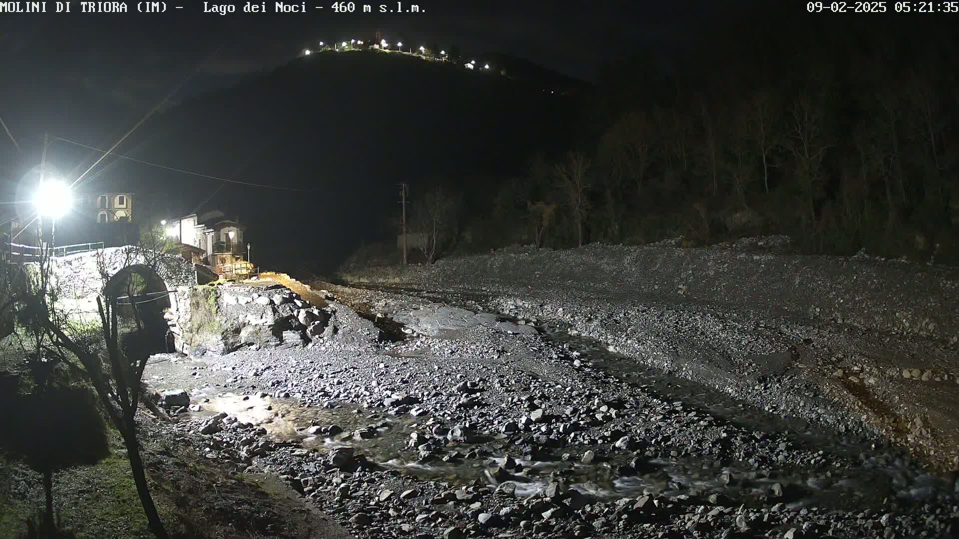 A rocky, shallow stream flows under a stone arch bridge at night, with lights illuminating the scene and a hilltop visible in the distance.