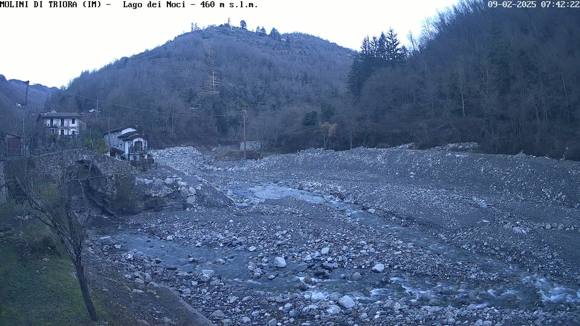 A rocky, shallow river flows through a barren riverbed beneath a hillside covered in leafless trees, under an overcast sky.