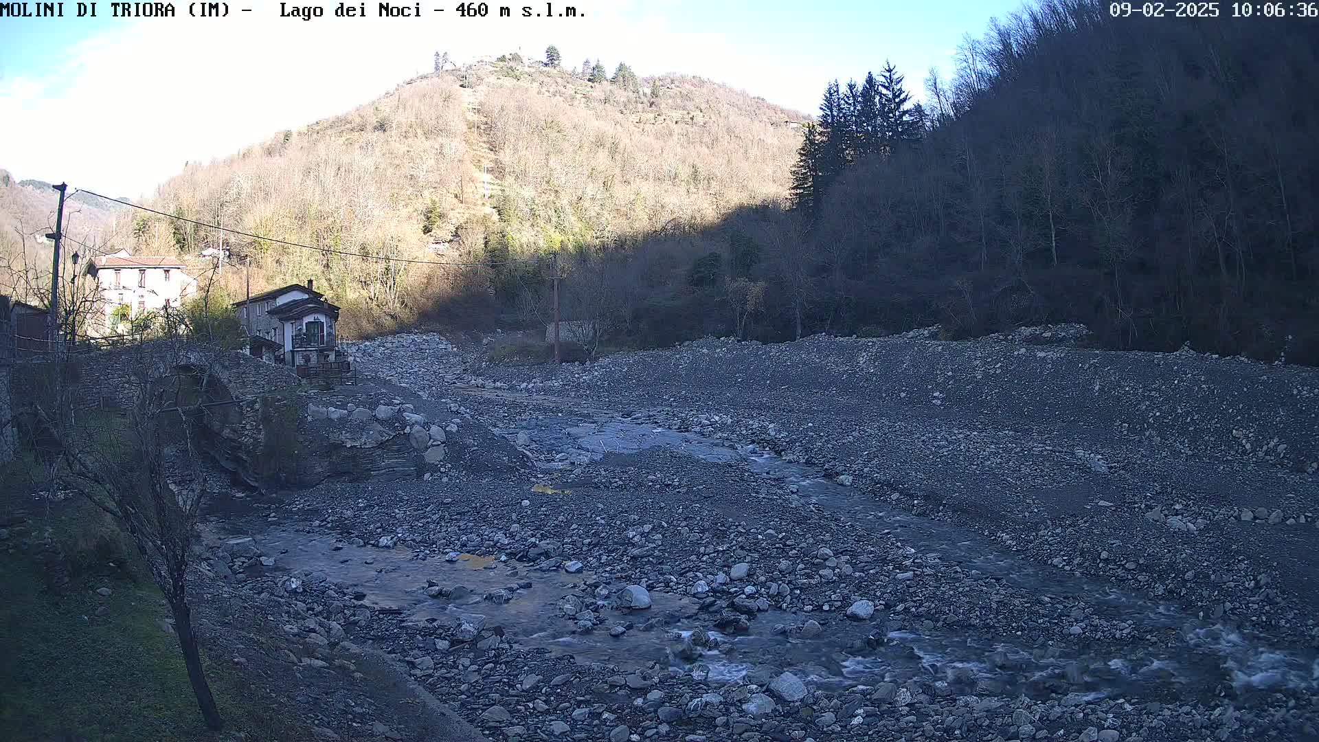 A rocky riverbed with a small stream flows past buildings and a bridge under a clear, sunny sky, nestled in a valley between leafless hillsides.