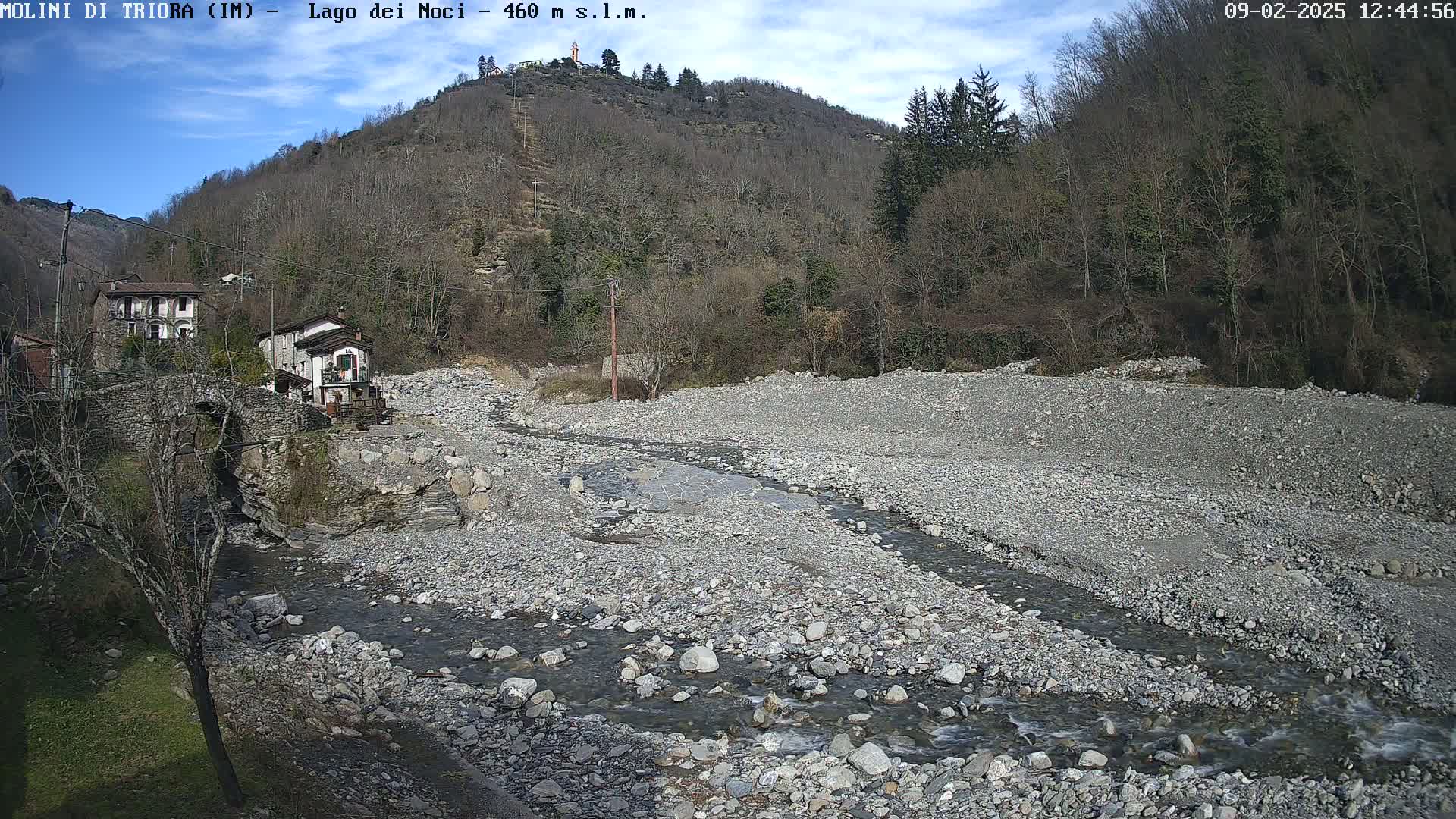 A rocky, dry riverbed with a small stream flows through it, past a stone bridge and buildings nestled in a hillside covered with leafless trees under a mostly clear sky.
